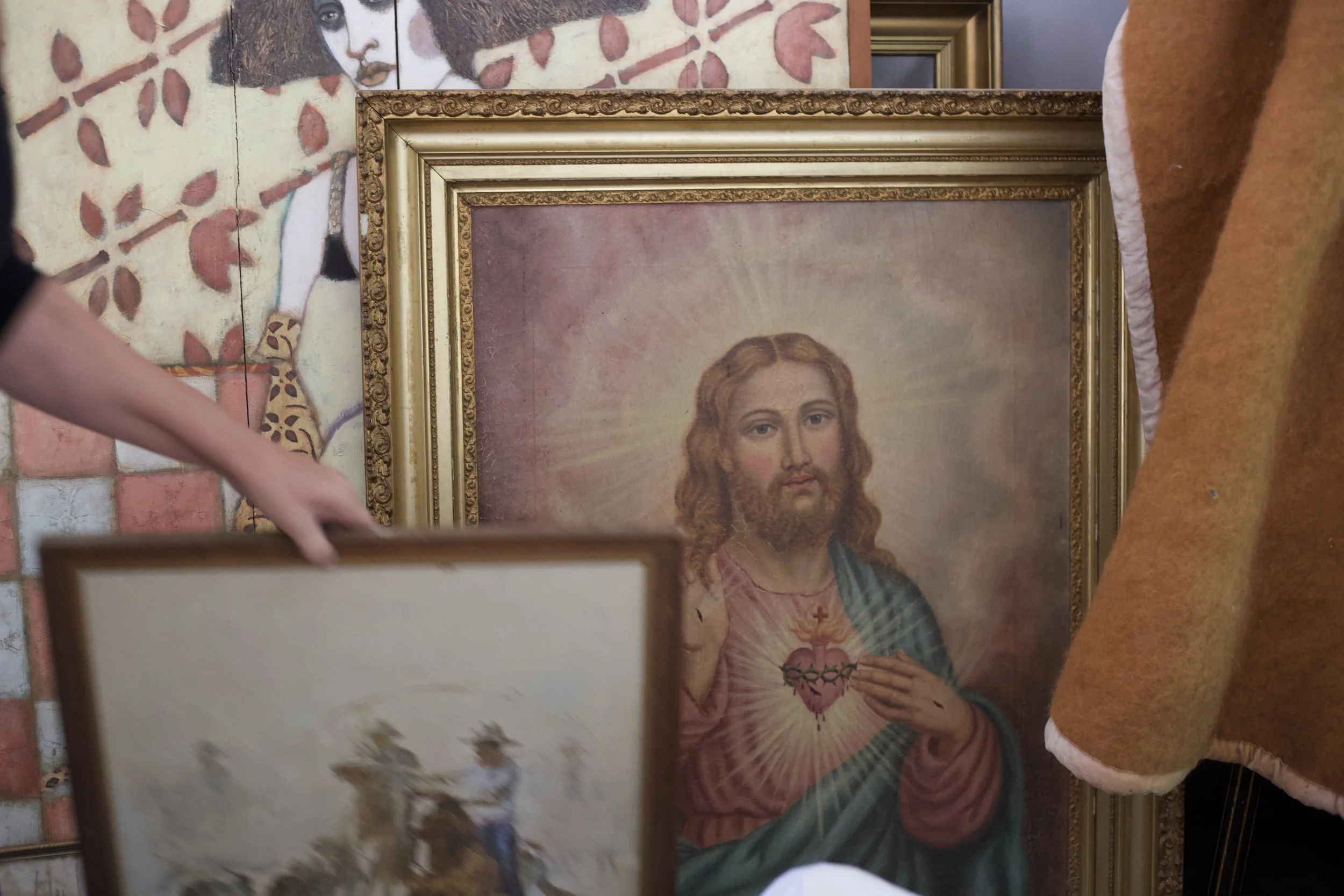 A framed religious painting of Jesus Christ with hand over heart, surrounded by several other framed artwork and paintings, with a person's arm visible in the foreground.