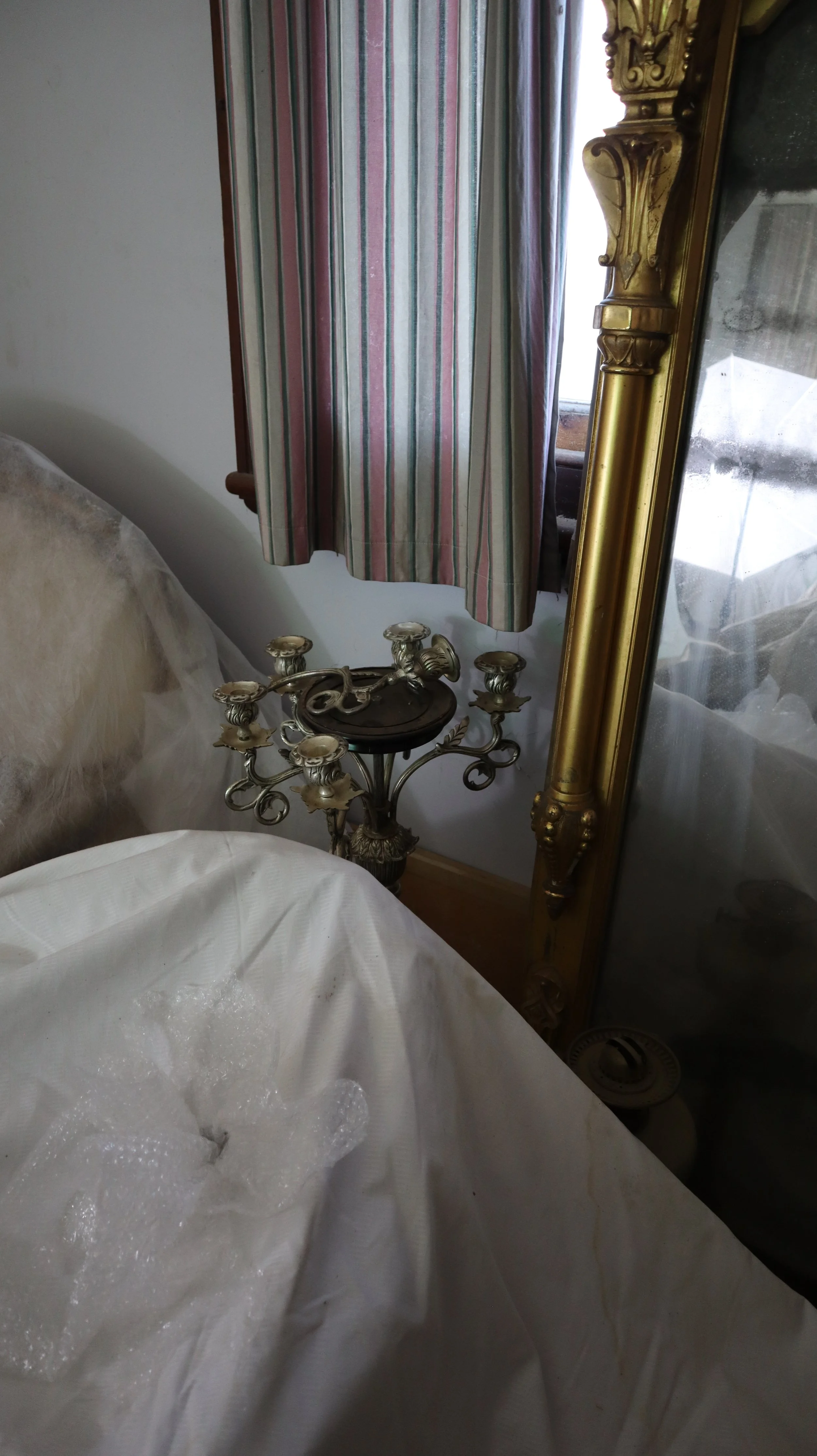 Close-up of a large ornate mirror with gold trim leaning against the wall, a window with striped curtains, and a small decorative metal stand with candles.