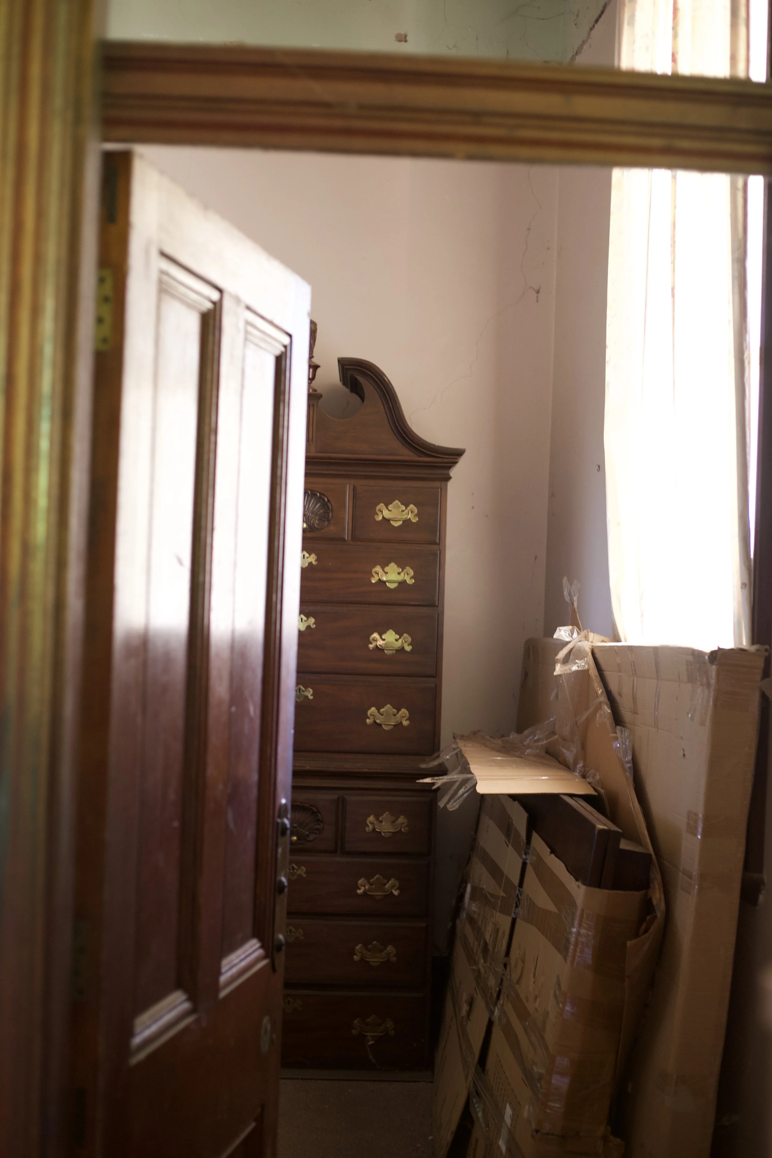 A view of a cluttered room with wooden furniture and cardboard boxes, visible through a door frame. The room has a window with light-colored curtains and high ceilings.