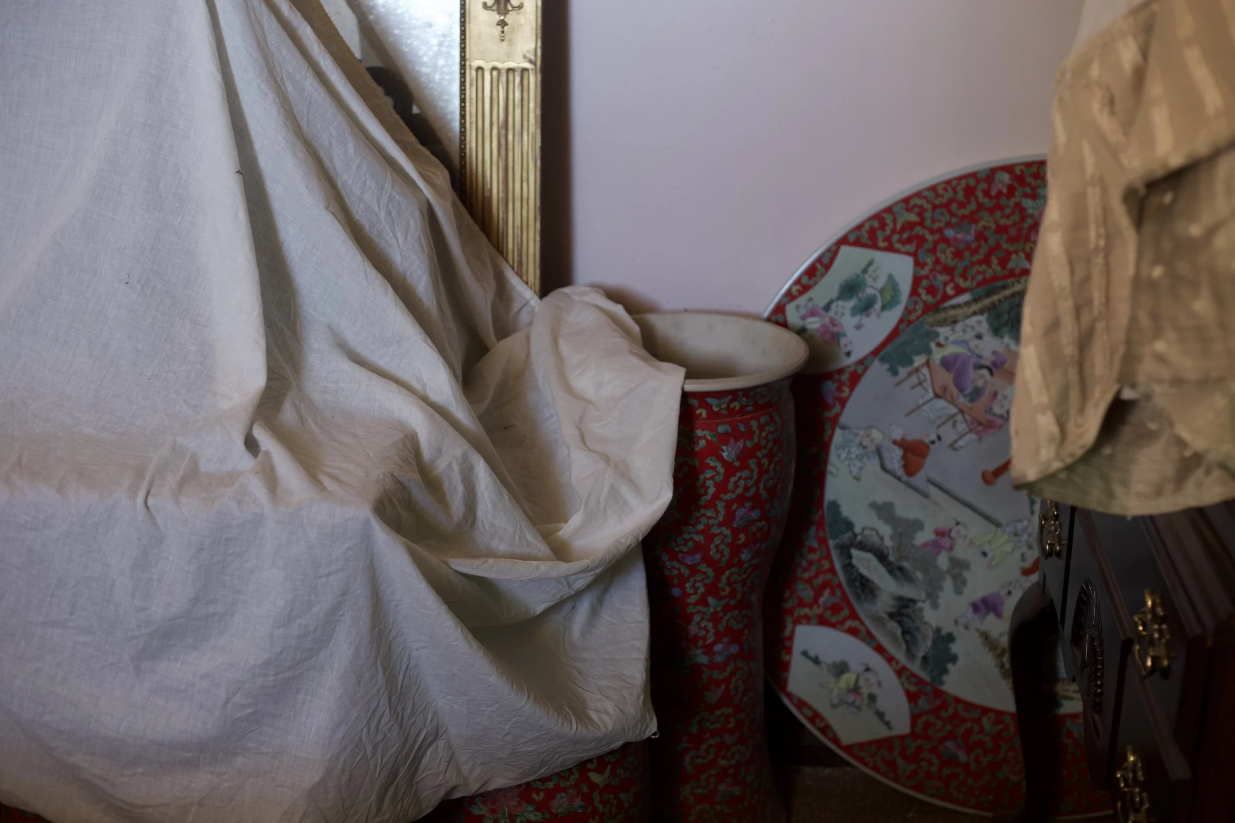 A cluttered room, with furniture draped in sheets. A Chinese ceramic item can be spotted.