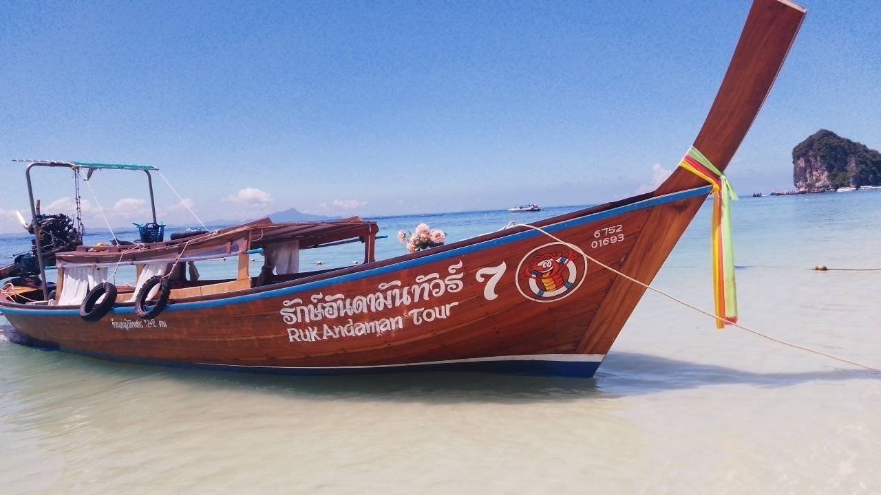 A traditional wooden long-tail boat floating on clear, shallow water near a sandy beach, with a rocky island and blue sky in the background.