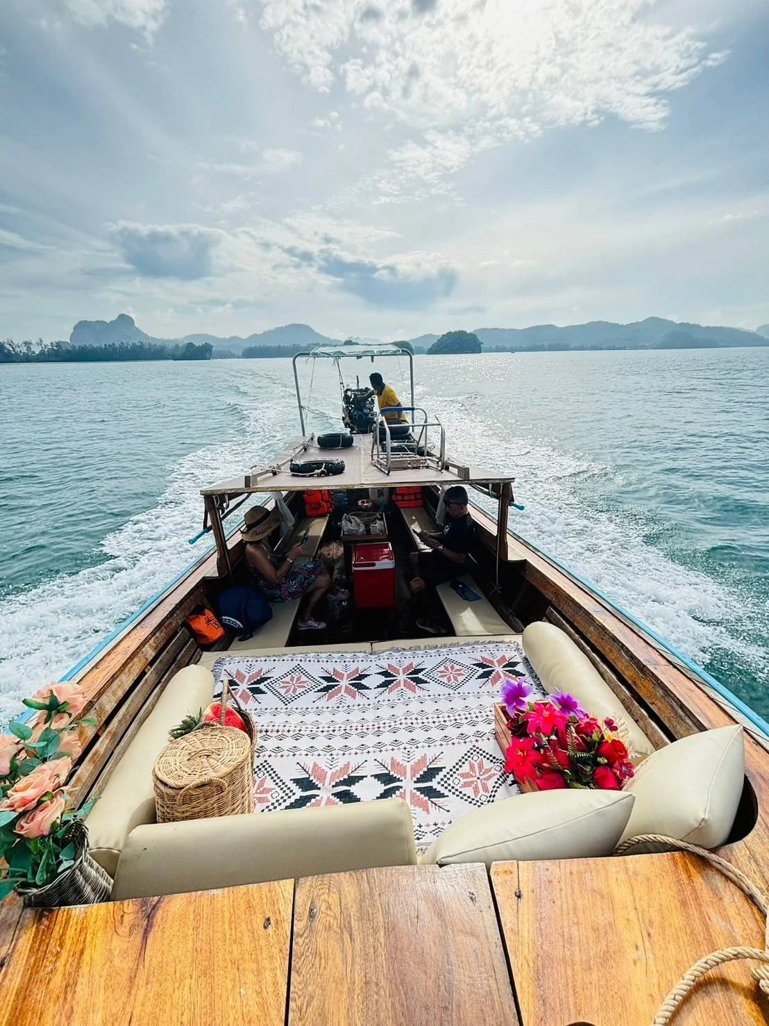 A boat with chairs, cushions, and flowers on the deck is moving on a body of water, with distant mountains and cloudy sky in the background.