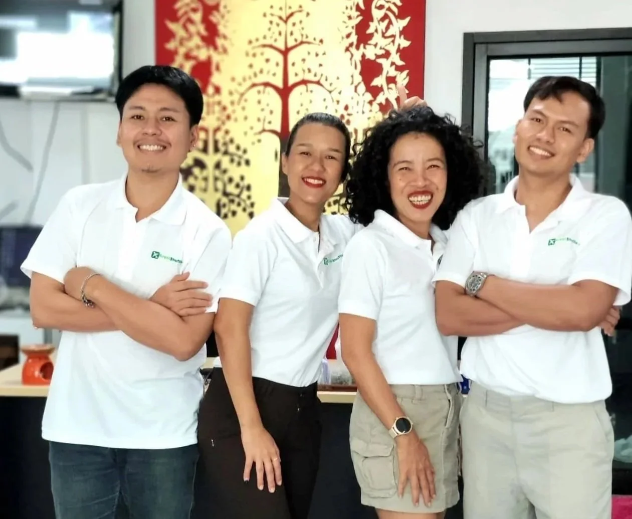 Four smiling people standing indoors in front of a decorative wall, wearing white polo shirts with a logo, appearing cheerful and confident.