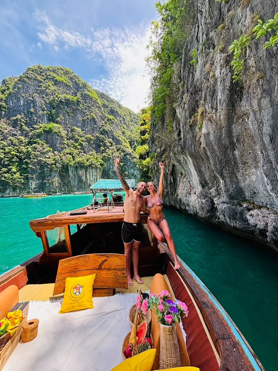 A man and woman in swimsuits on a boat with a turquoise body of water, surrounded by tall, rocky, green islands, under a partly cloudy sky.