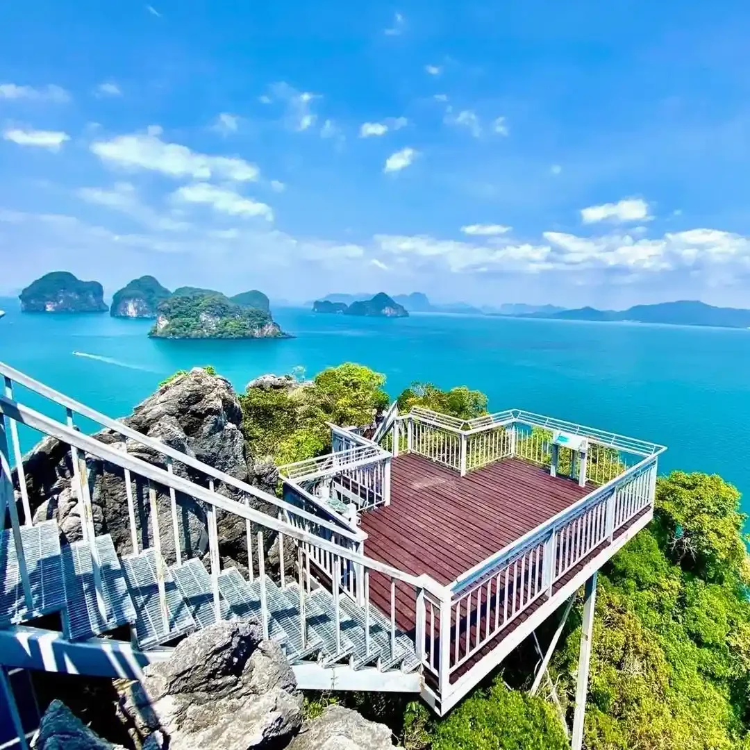 A scenic overlook balcony with white railings and wooden flooring on a rocky hilltop, overlooking a turquoise sea with small islands and a partly cloudy blue sky.