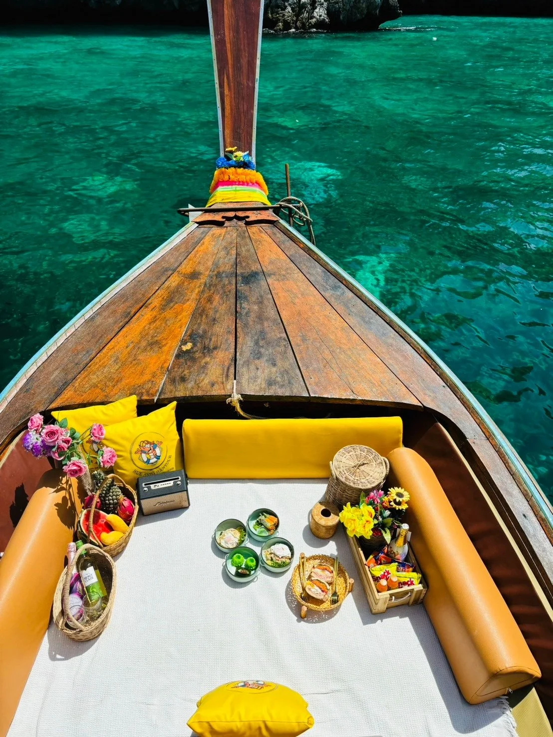 A boat's bow with a white mat, yellow cushions, and various items including flowers, food, and drinks, cruising on clear turquoise water.