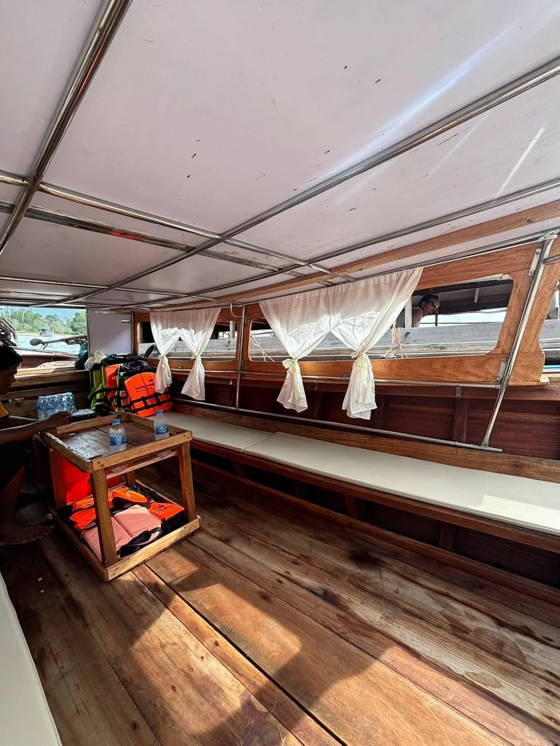 Interior of a wooden boat with a white canopy, windows with white curtains, and a long bench seat along the side. There are some water bottles and life jackets on a small wooden table.