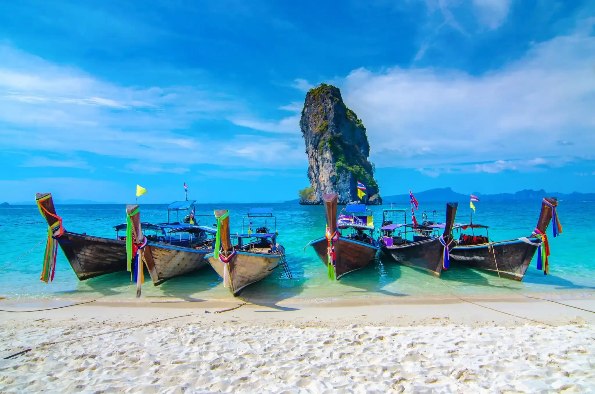 Traditional Thai longtail boats with colorful ribbons tied to the bows parked on a sandy beach with clear turquoise water, large limestone karst island in the background, and a blue sky with scattered clouds.
