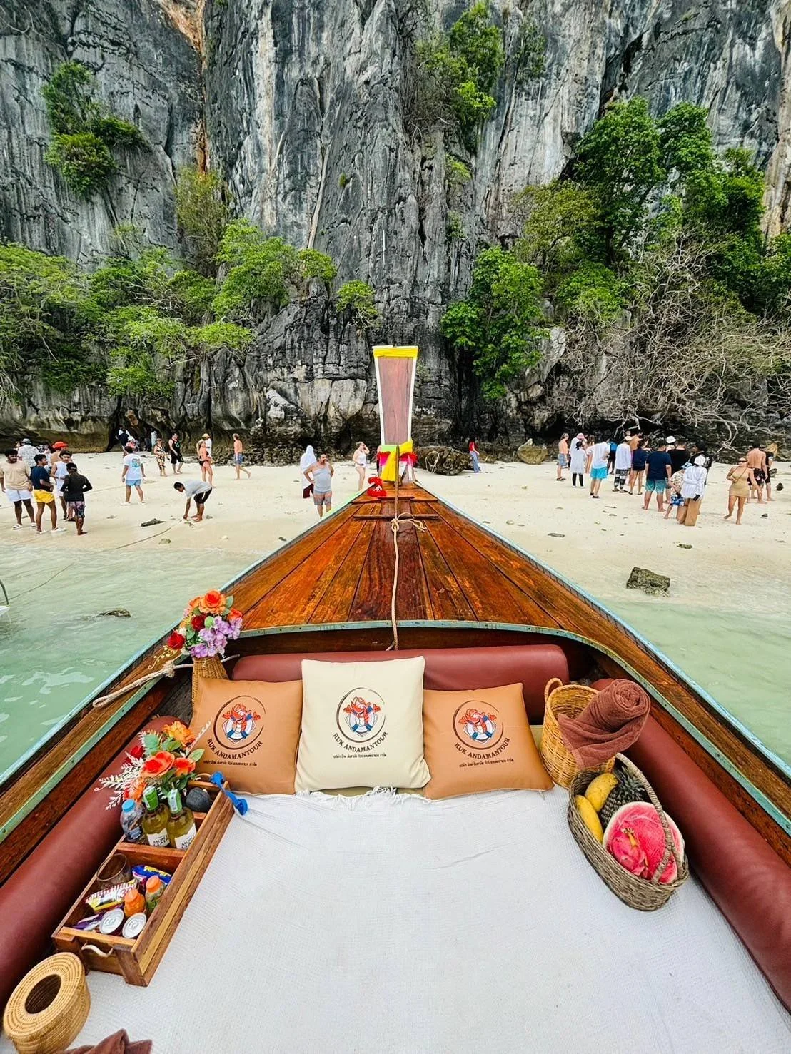 View from a boat with a wooden deck, colorful pillows, and water bottles, facing a beach with rocks and costly cliffs, and people walking and swimming.