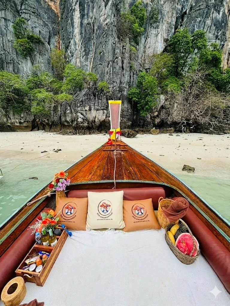 View from the front of a wooden boat docked near a sandy beach with rocky cliffs and green trees in the background. Inside the boat, there are pillows, a basket with rolled towels, a tray with supplies, and decorative items.