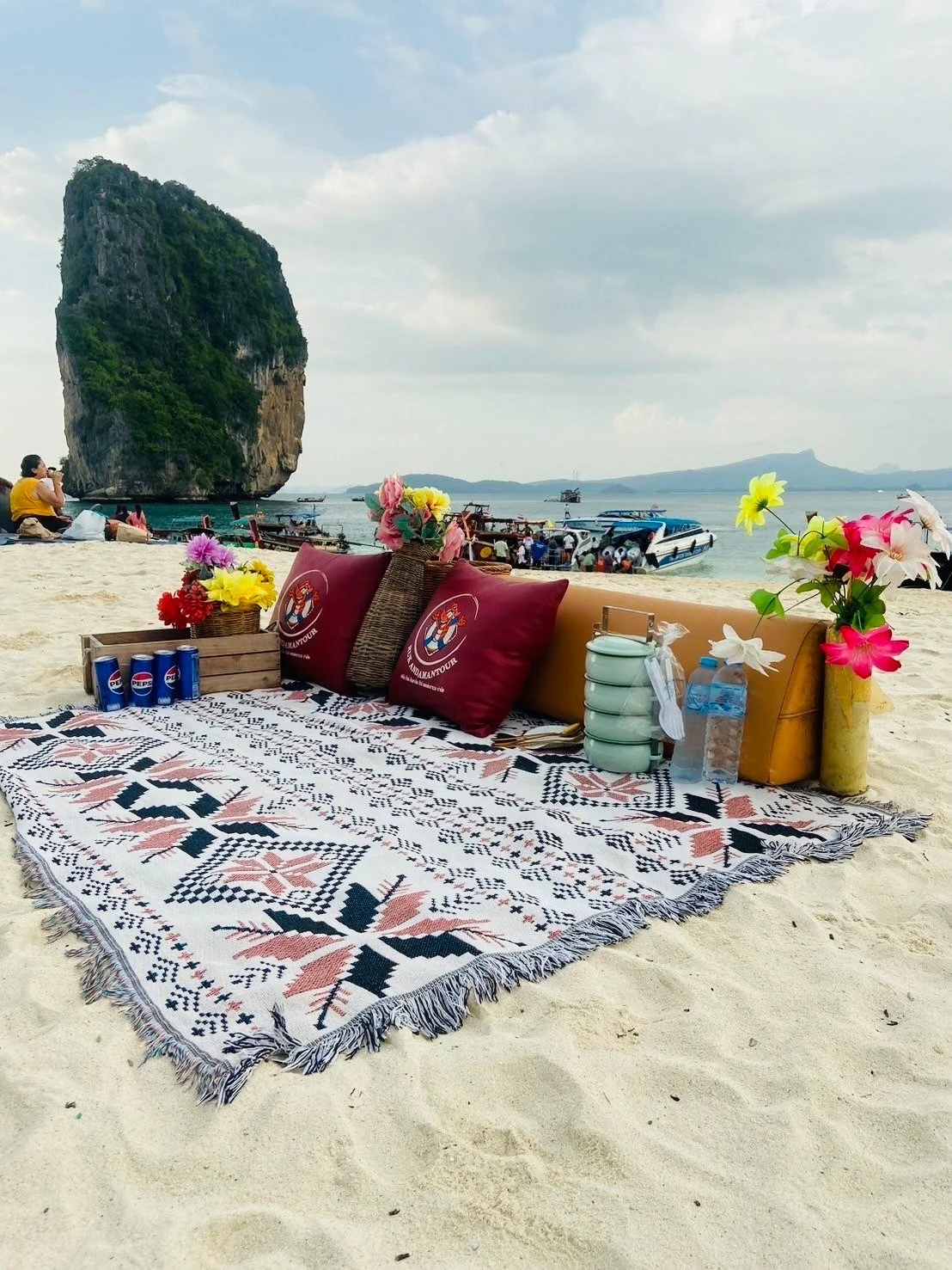 A beach scene with a patterned blanket, pillows, flowers, and drinks in the foreground. In the background, boats are docked near large rock formations and a person is sitting on the sand taking a photo.