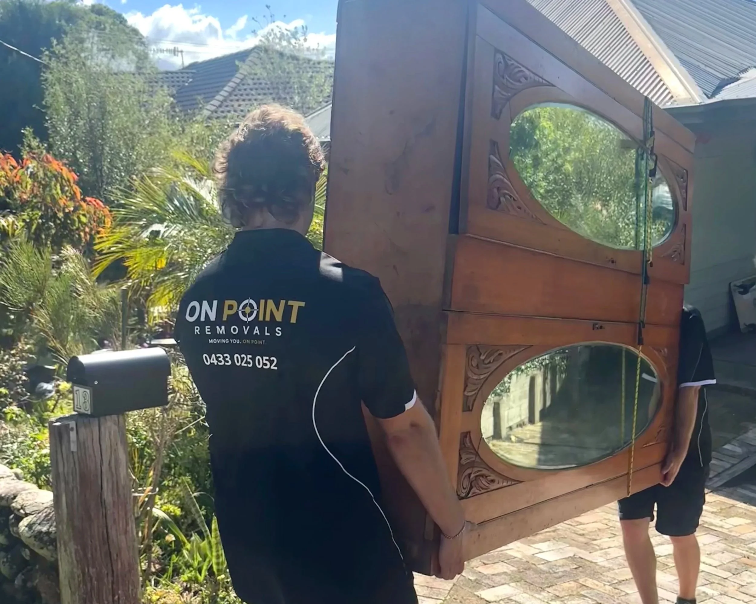 Person wearing a black shirt with white text, helping move a large wooden piece with carved designs, outdoors near greenery and houses.
