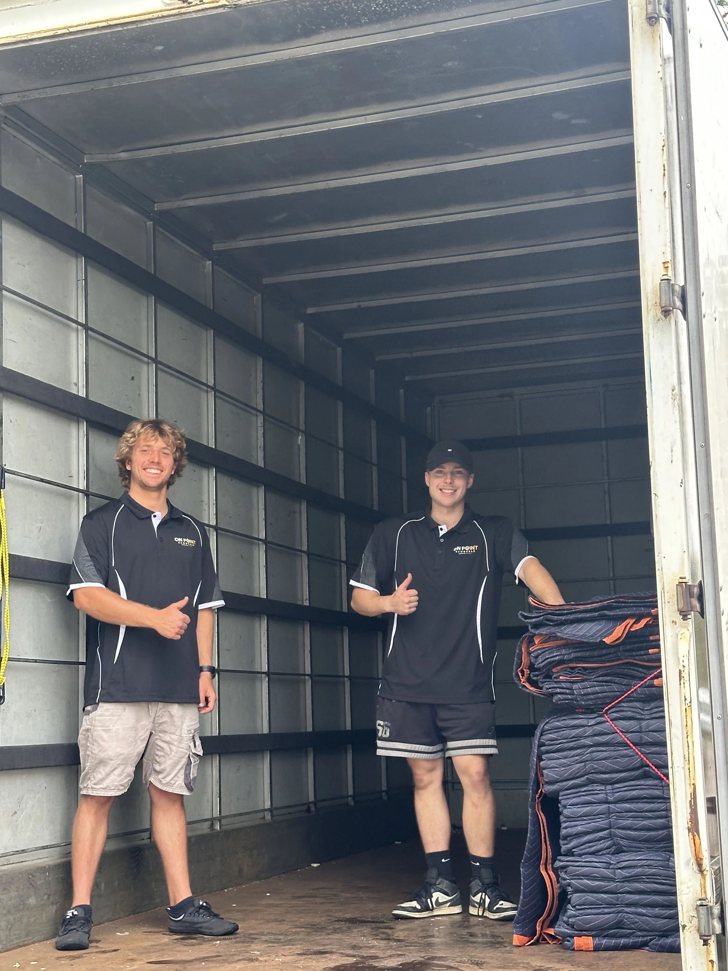 Three young men standing inside the open back of a moving truck, smiling and posing for the photo. One is giving a thumbs-up, wearing shorts and a black polo shirt with a logo, and the other two are also dressed in black and standing next to a pile of folded blankets or pads.