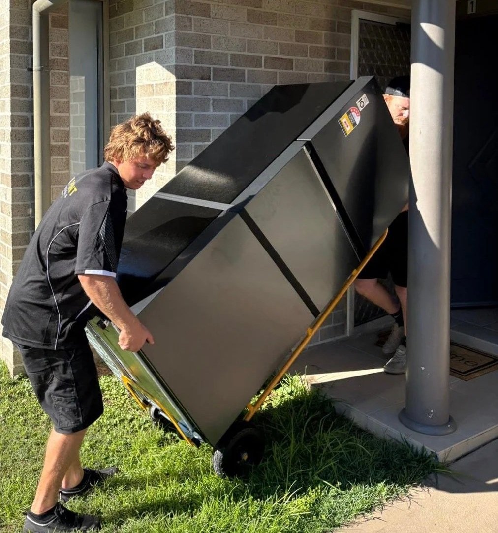 Two young men are moving a large, black, commercial refrigerator on a hand truck outside a building with brick walls. One man is pushing the appliance while the other is helping from behind.