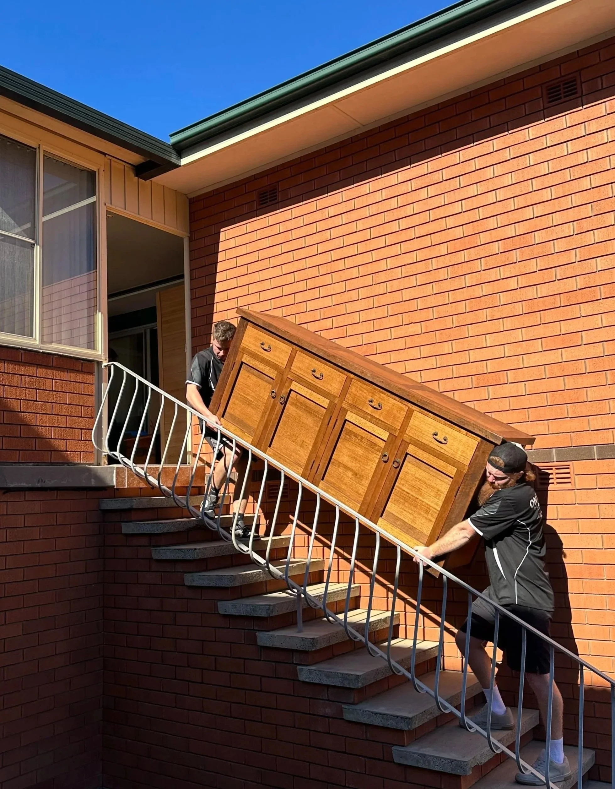 Two men are carrying a wooden dresser up stairs on an outdoor brick staircase. The house has brick walls and a window, with a clear blue sky above.