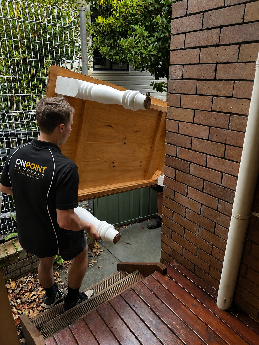 A person in a black ON POINT REMOVALS shirt carrying a large white plastic pipe on a wooden board, outside near a brick wall and on a deck with steps, with green foliage in the background.