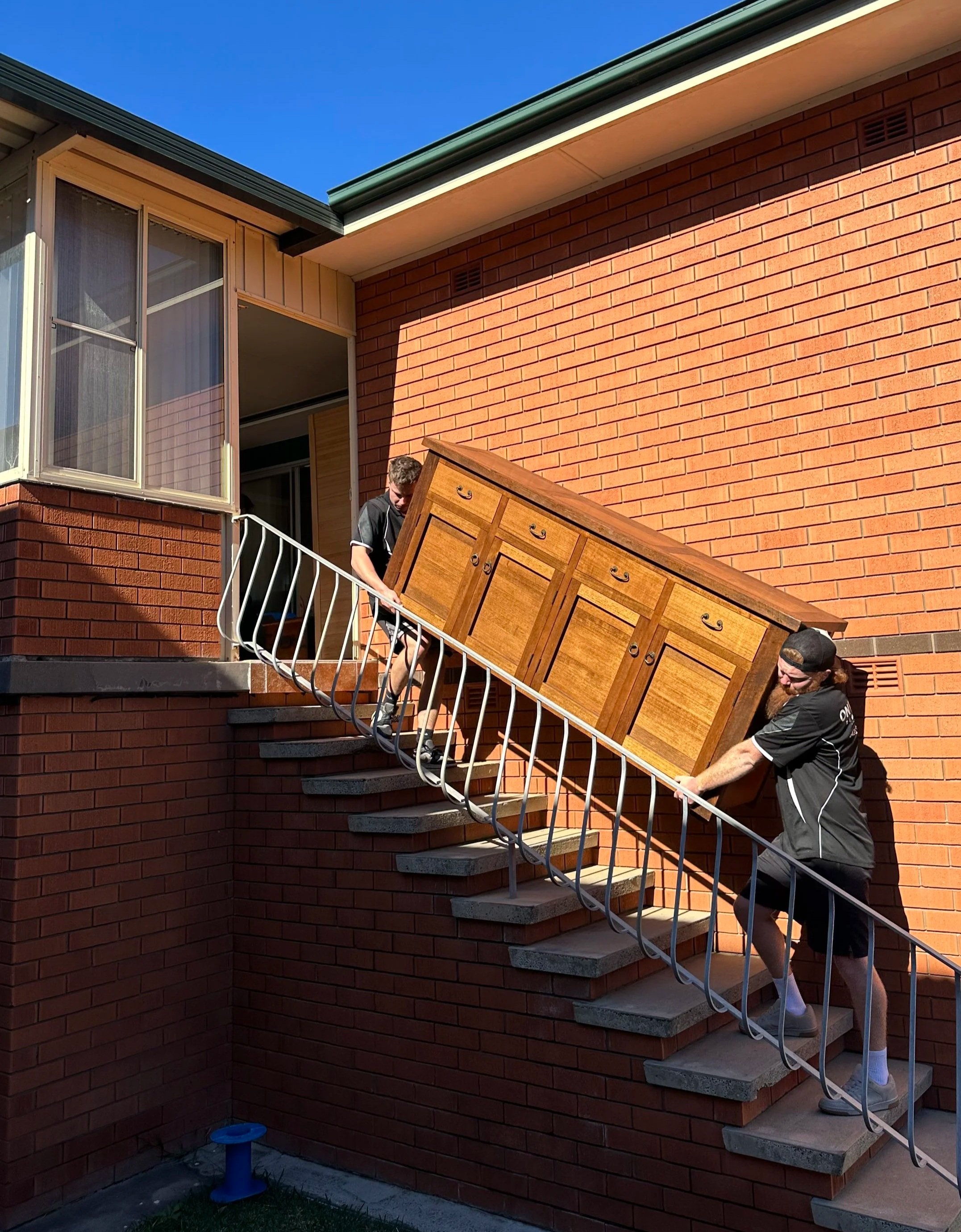 Two men carry a large wooden cabinet down outdoor concrete stairs attached to a red brick house on a bright sunny day.