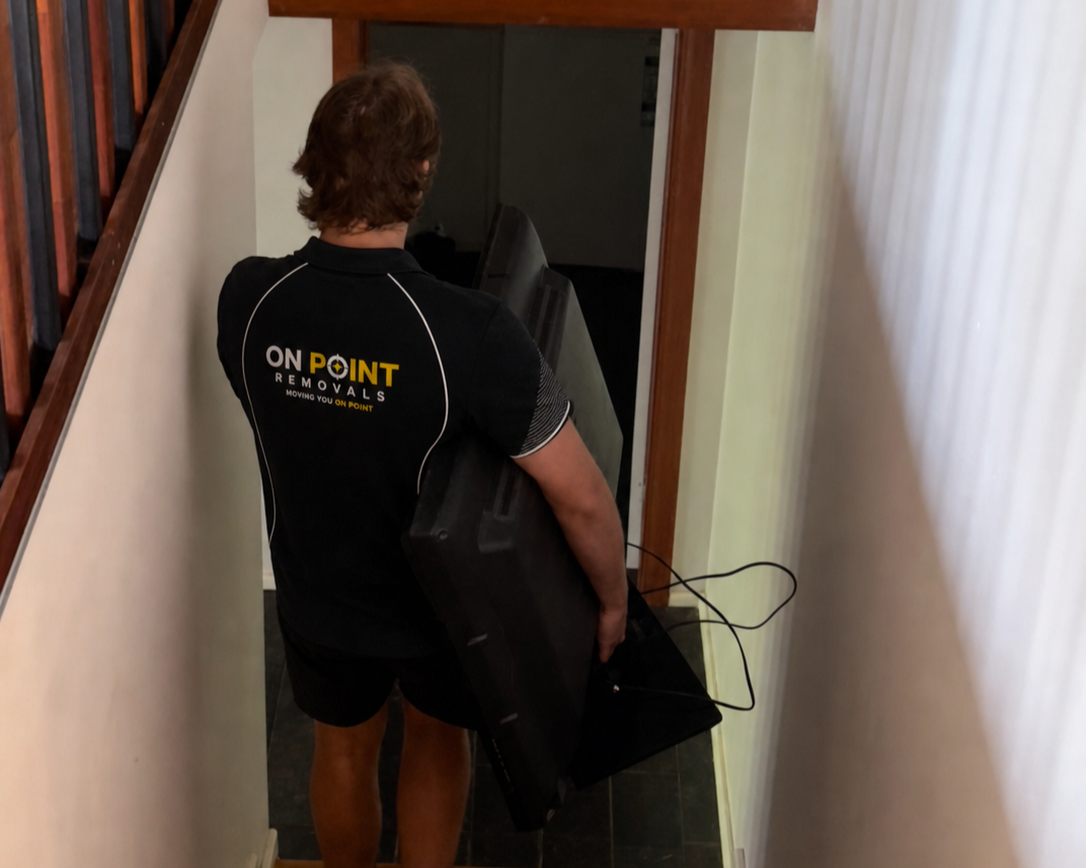 A man wearing a black ON POINT REMOVALS shirt carrying a large flat-screen TV down a staircase.