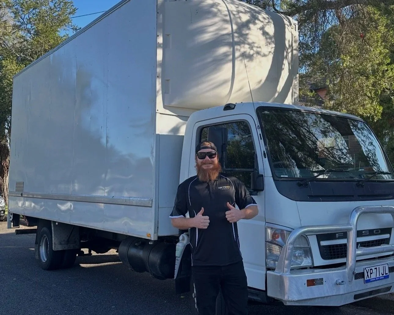 A man with a beard, sunglasses, and a black shirt standing in front of a white box truck, giving a thumbs-up gesture outdoors during daytime.