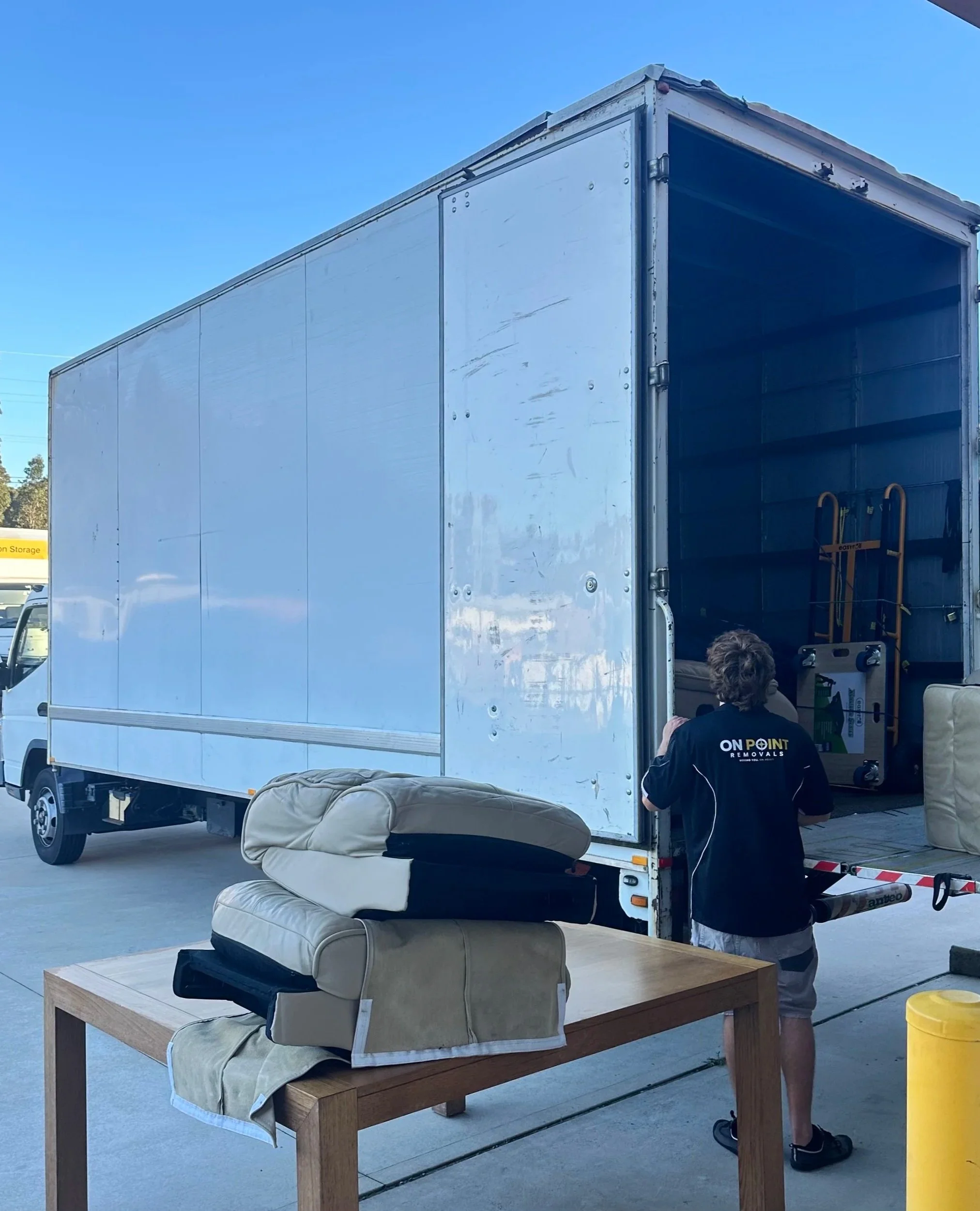 A white moving truck with its rear door open, inside which there is a yellow hand truck. In front of the truck, a person with short curly hair wearing a black shirt with the words 'ONPOINT REMOVALS' on the back, khaki shorts, and black shoes is standing. There is a sofa with a beige cover and a wooden table with another sofa and a yellow bucket nearby, on a concrete surface outdoors under a clear blue sky.