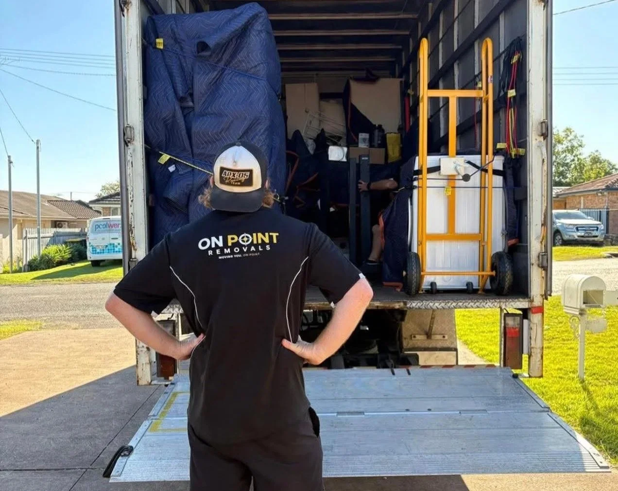 Man wearing a black shirt with 'ONPOINT REMOVALS' logo on back, standing with hands on hips in front of an open moving truck filled with packed belongings, including furniture and a yellow hand truck, on a driveway in a residential neighborhood.