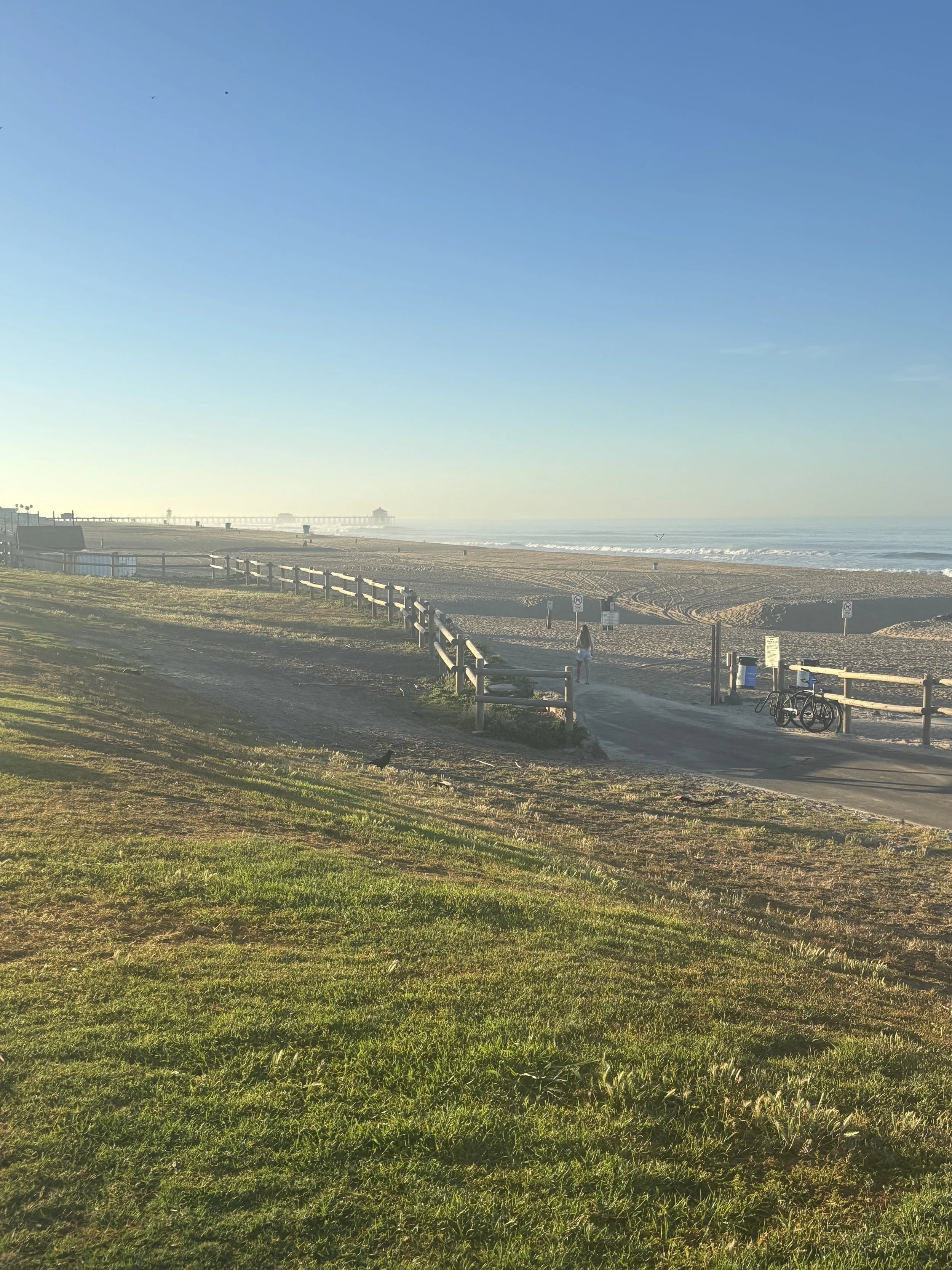 A sandy beach with a pier in the distance, a person walking along the shoreline, and bicycles parked near a fenced area with some trees and a grassy area in the foreground.