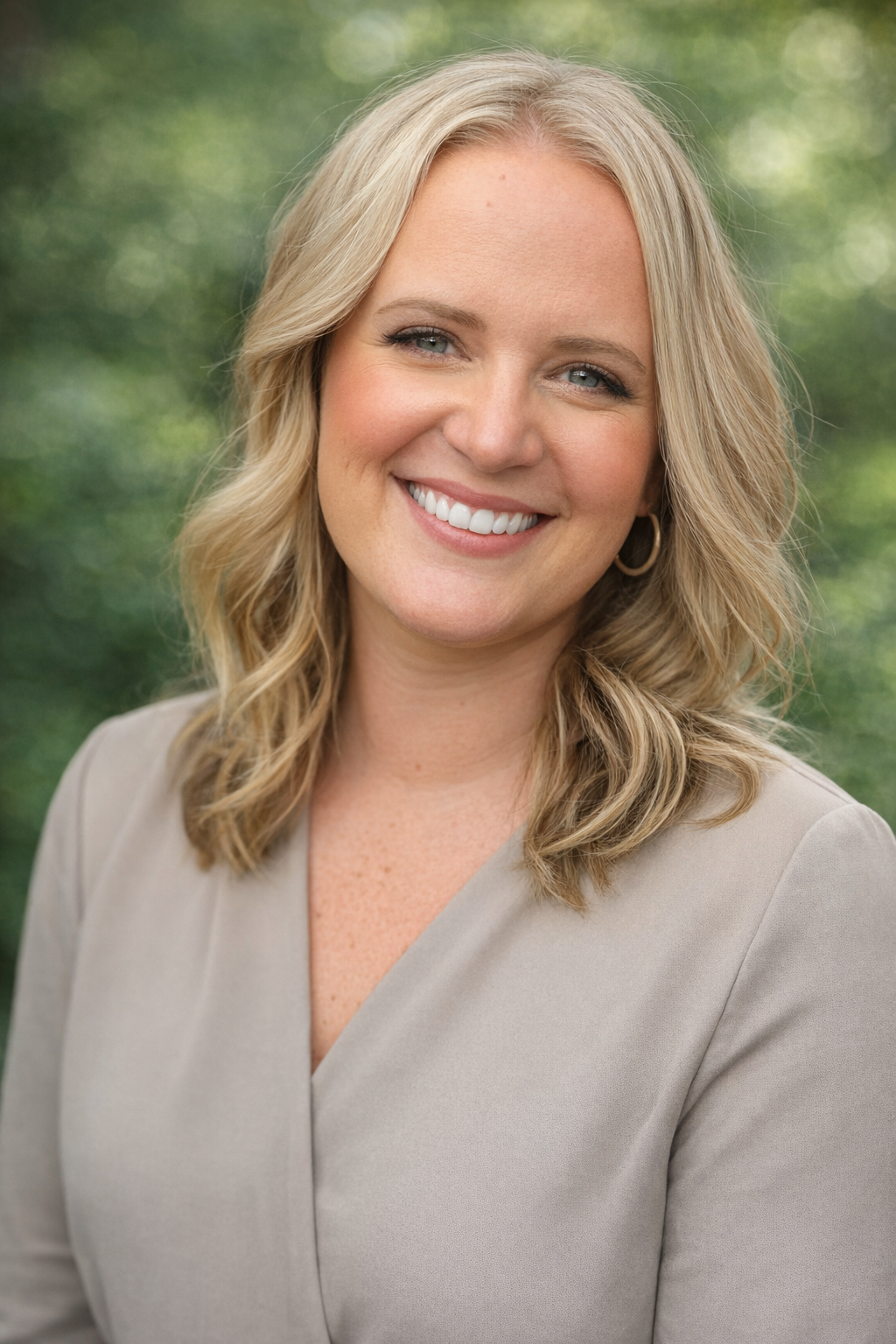 A woman with blonde hair smiling outdoors, wearing a beige blazer and hoop earrings, with a blurred green natural background.