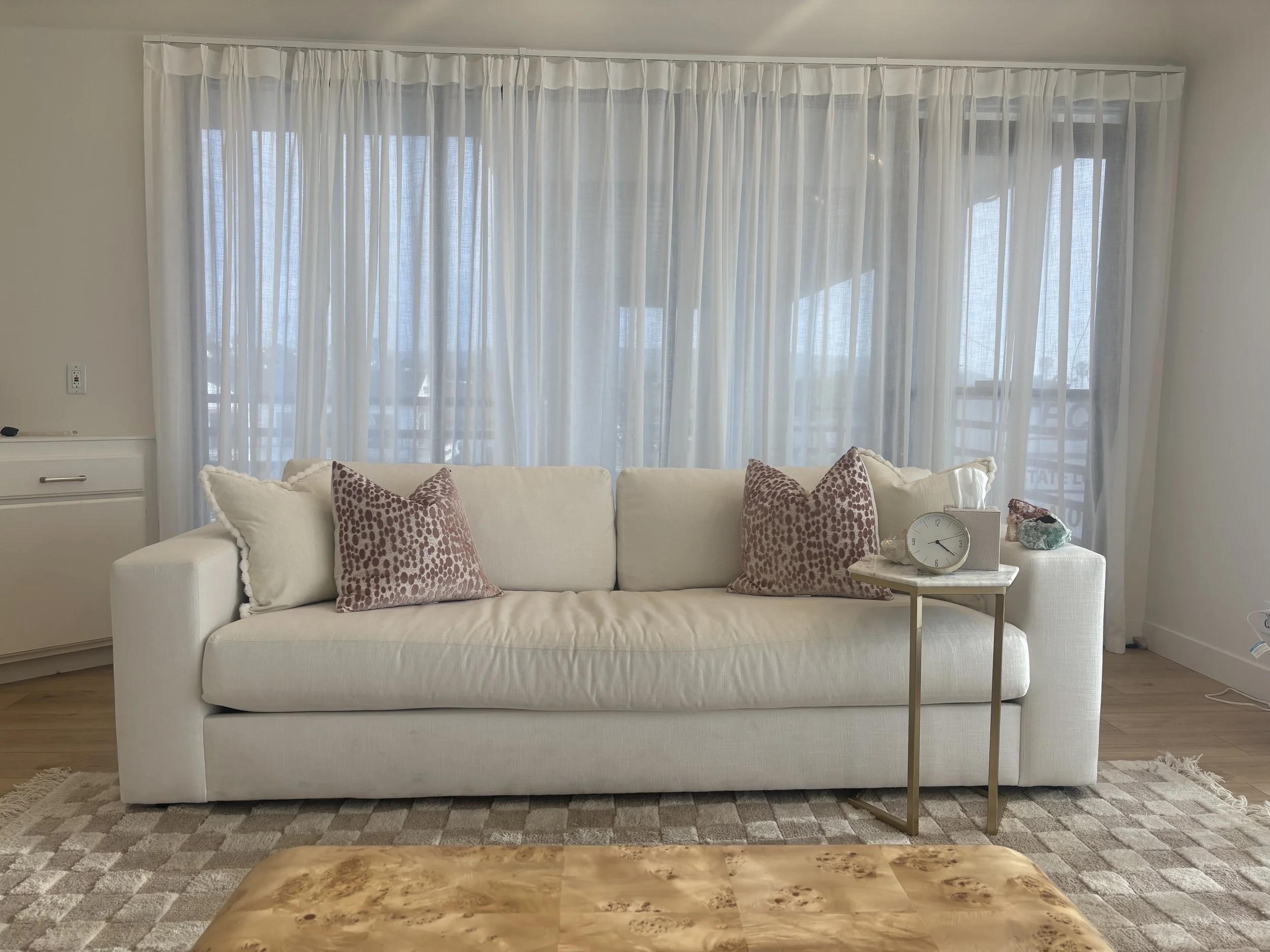 Living room with white sofa, patterned pink pillows, a marble side table with a clock and stones, behind large sheer curtains covering windows.