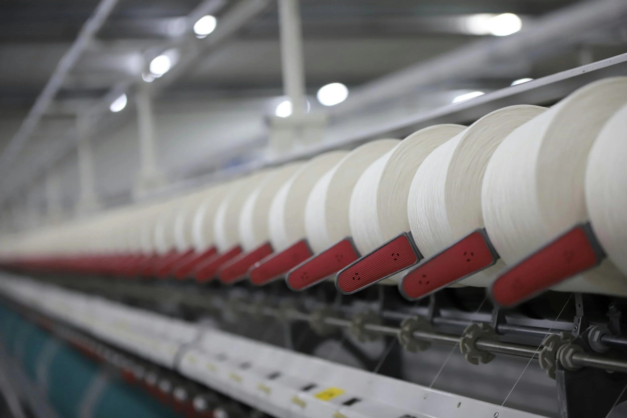 Close-up of a sewing machine's spools of white thread with red tensioners, arranged in a row in a manufacturing environment.