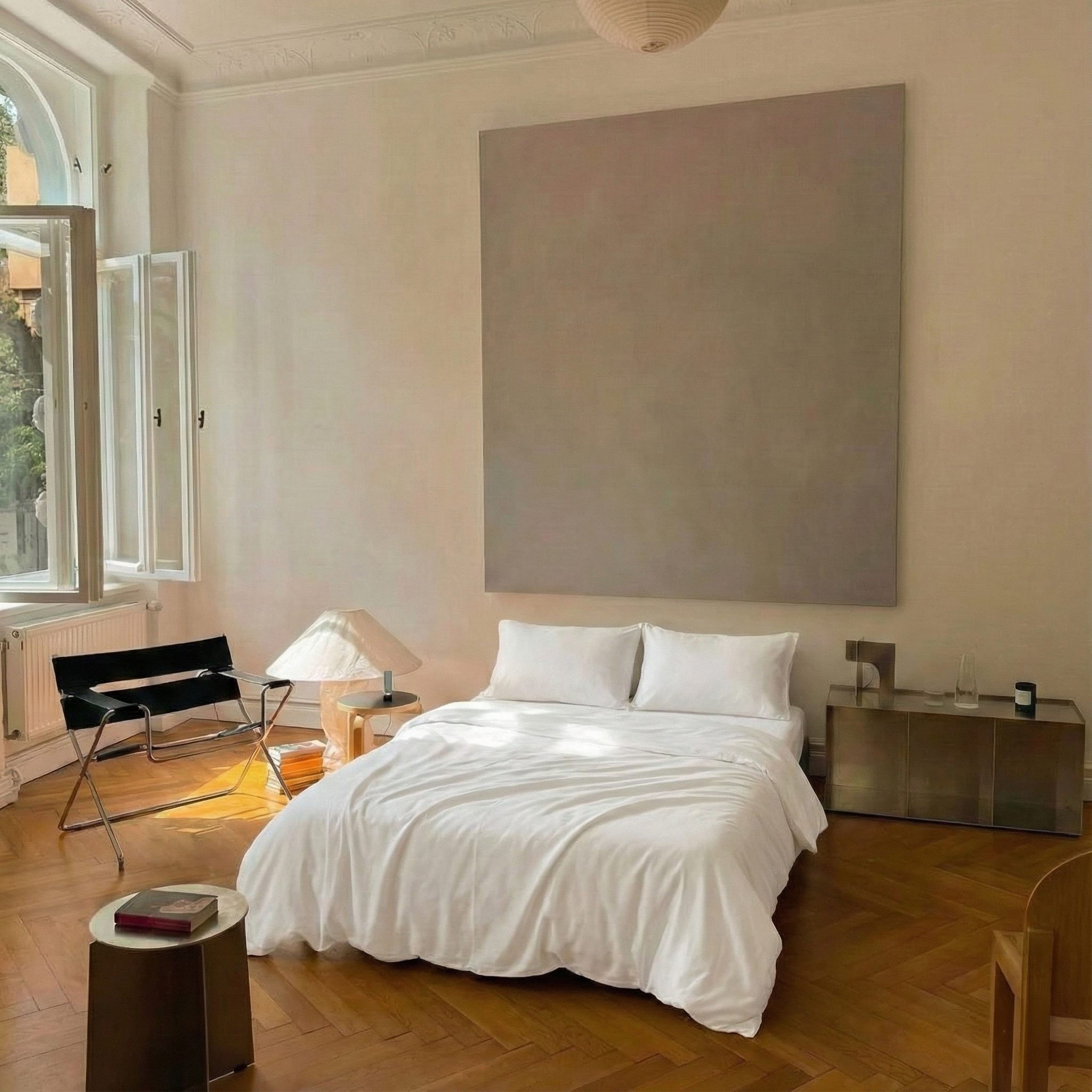 A bedroom with a large window on the left, an unmade bed with white sheets and pillows in the center, a nightstand with a lamp and a small table with a book on it, and a black lounge chair near the window. The room features wooden flooring and minimal decor.