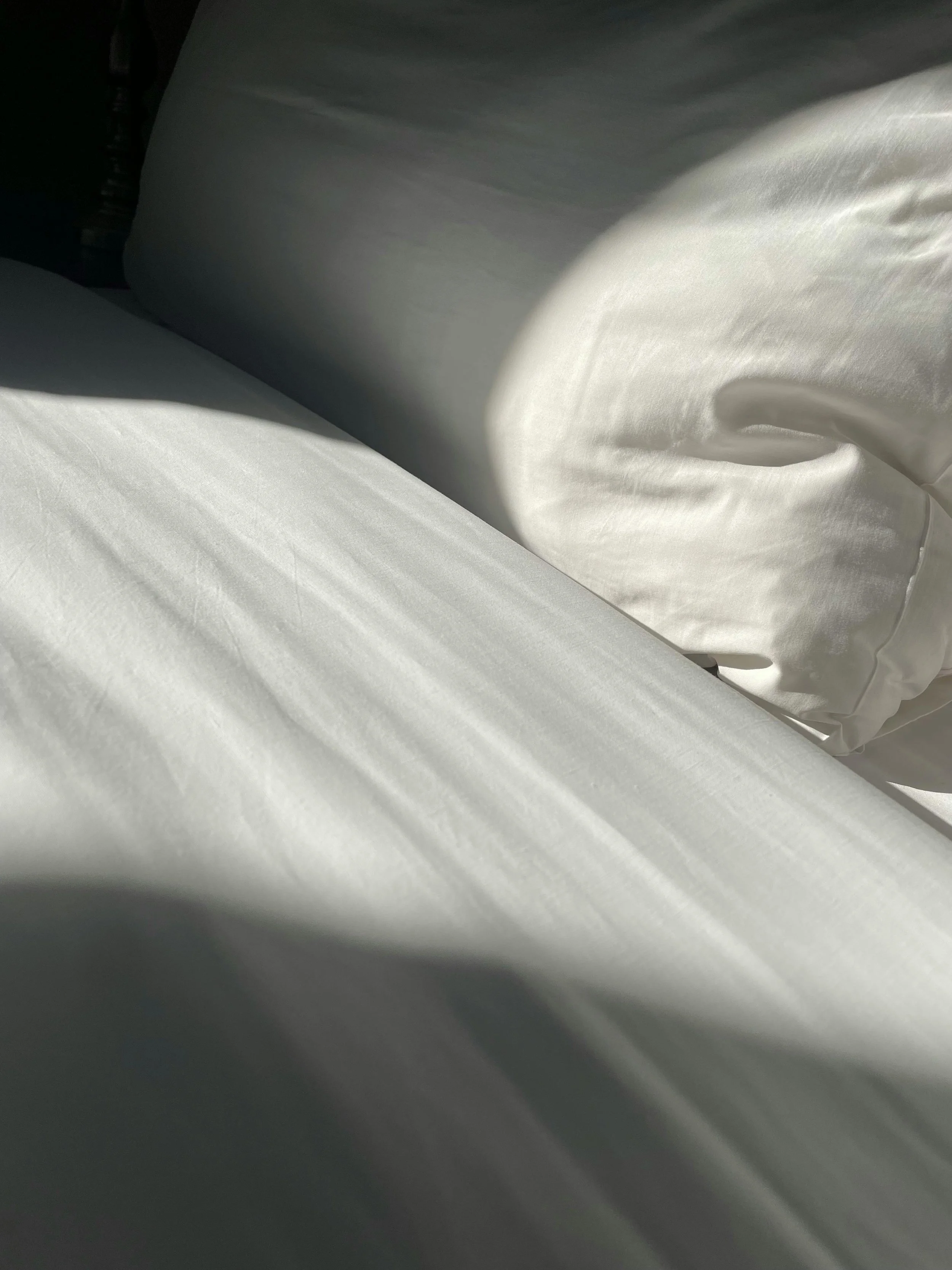 Close-up of white bed sheets and pillow with shadows cast by sunlight.