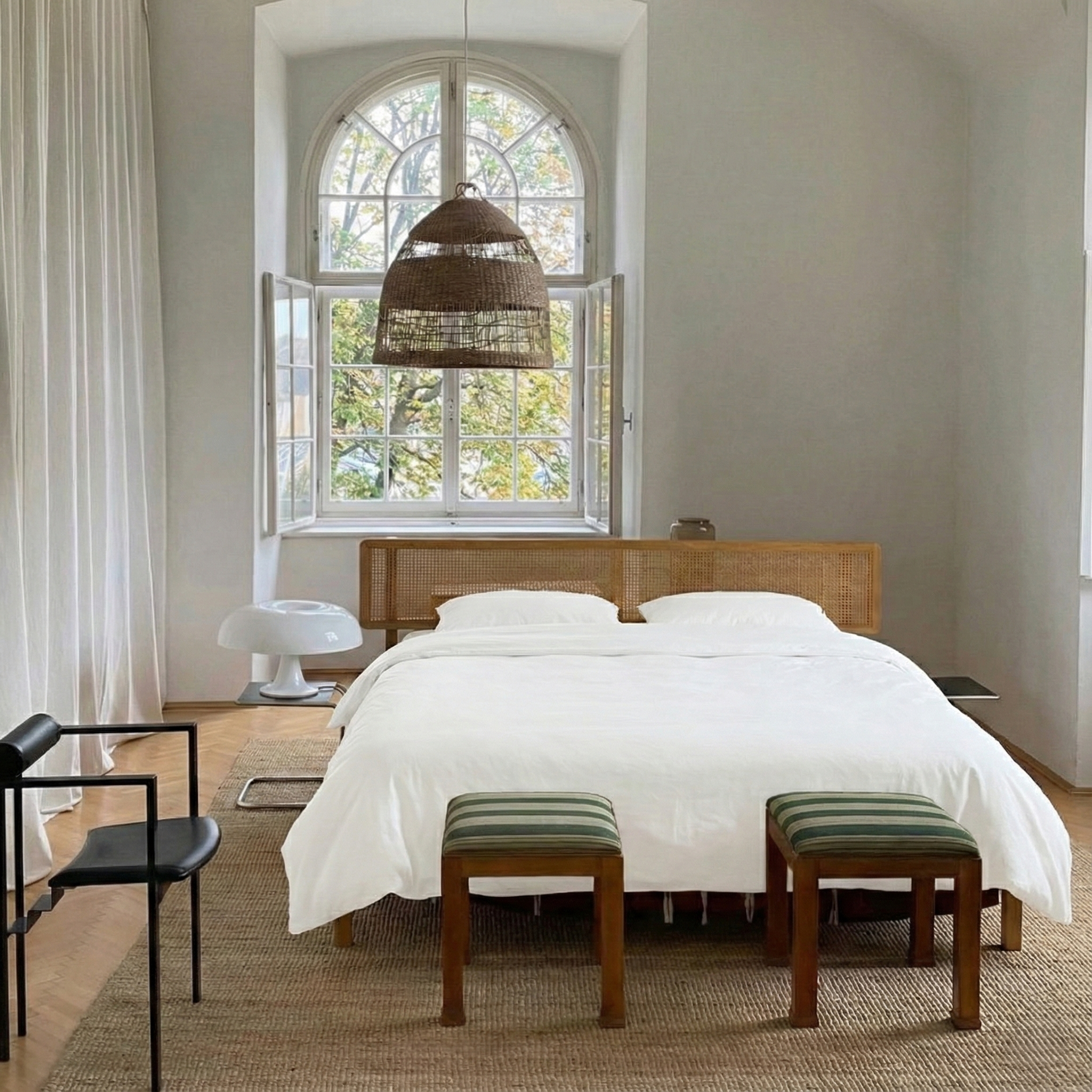 A minimalist bedroom with a large, white bed, two small striped stools at the foot of the bed, a round side table, a black chair, and large arched window with open shutters allowing natural light to fill the room.