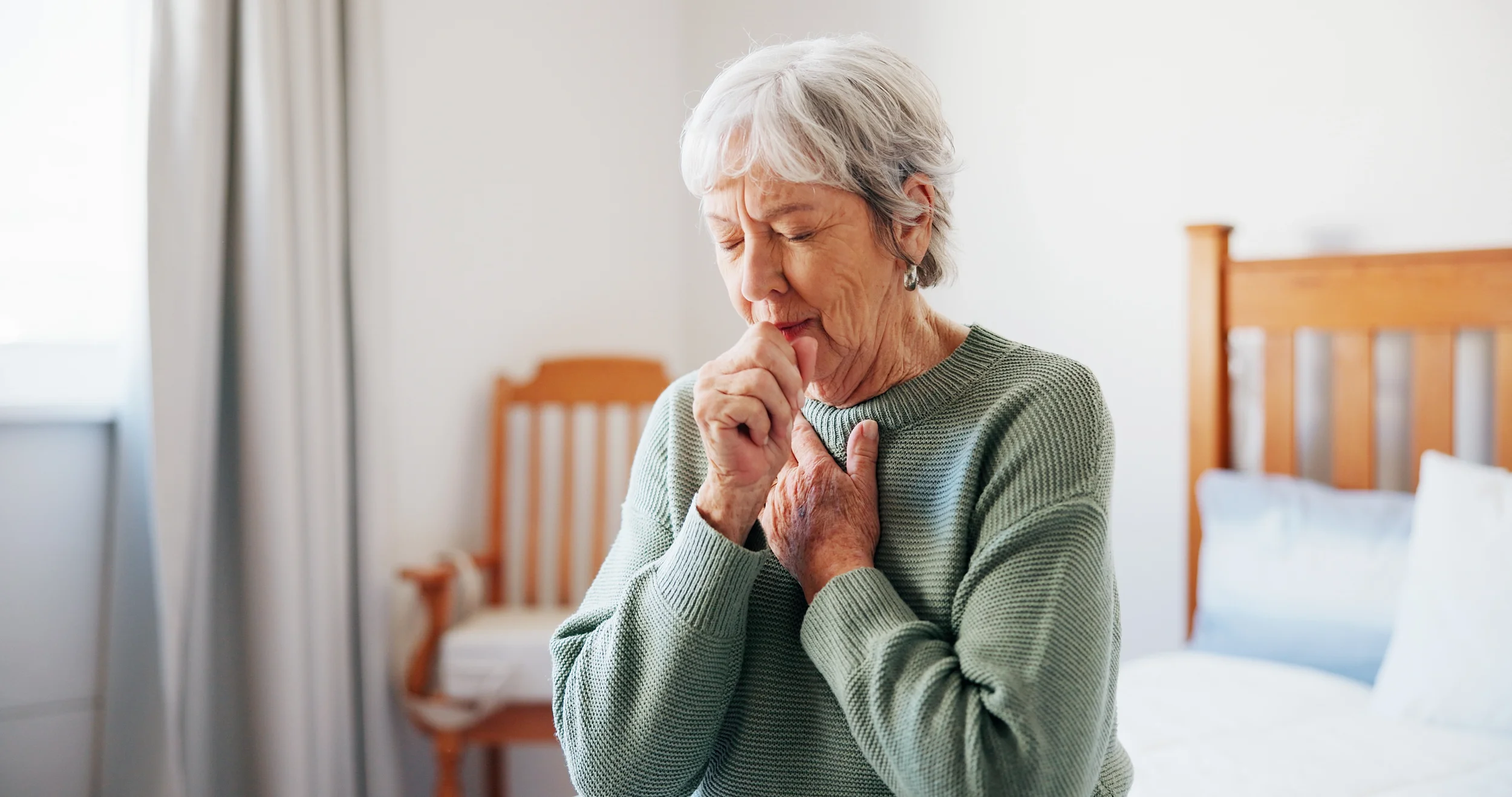 An elderly woman with gray hair and a green sweater appears to be distressed, clutching her chest with one hand and covering her mouth with the other, inside a bedroom with wooden furniture and light-colored curtains.