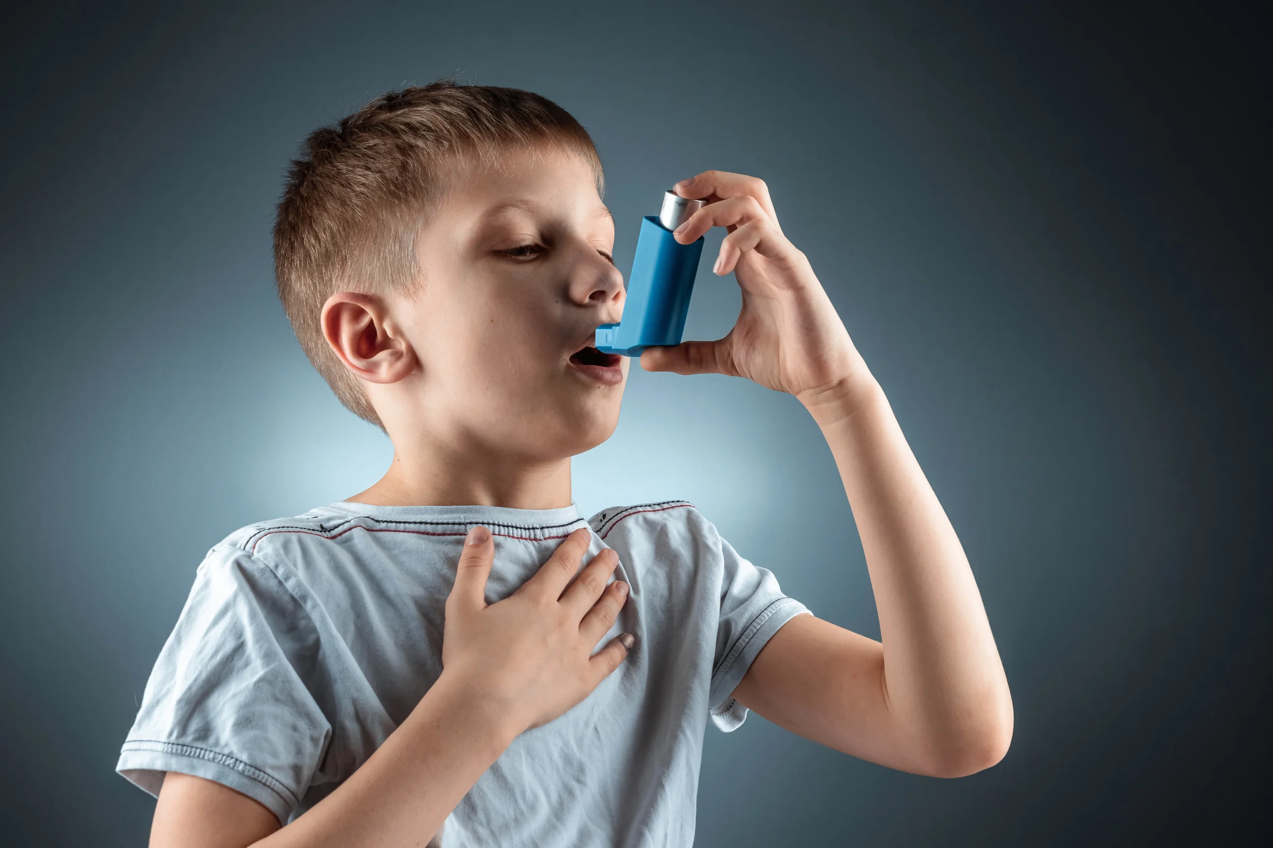 A young boy with short hair and a striped shirt is holding an inhaler near his mouth, with one hand on his chest, against a plain background.