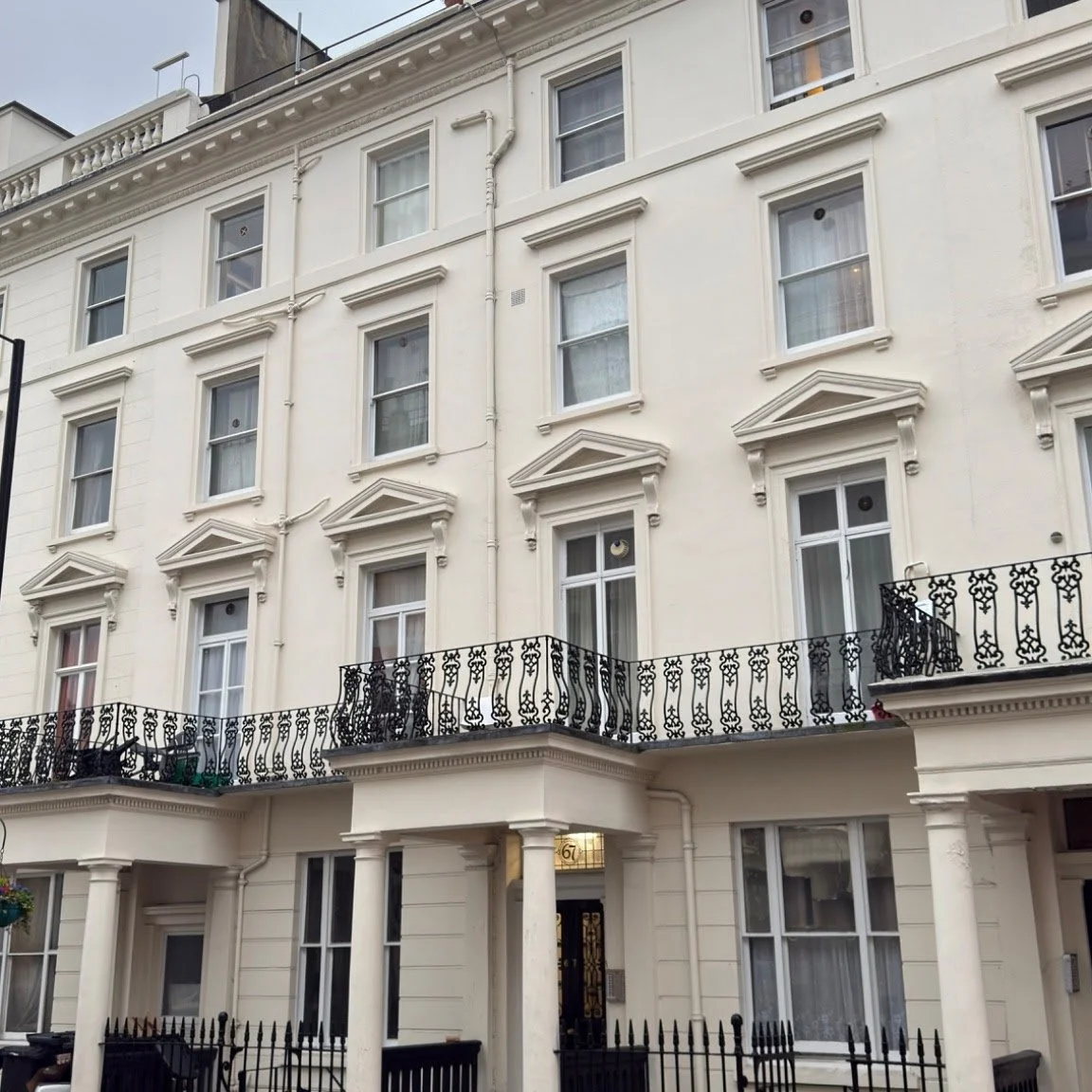 Four-story cream-colored building with white window trims, decorative molding, and wrought iron balconies. The building has a black iron fence and entryway with column supports.