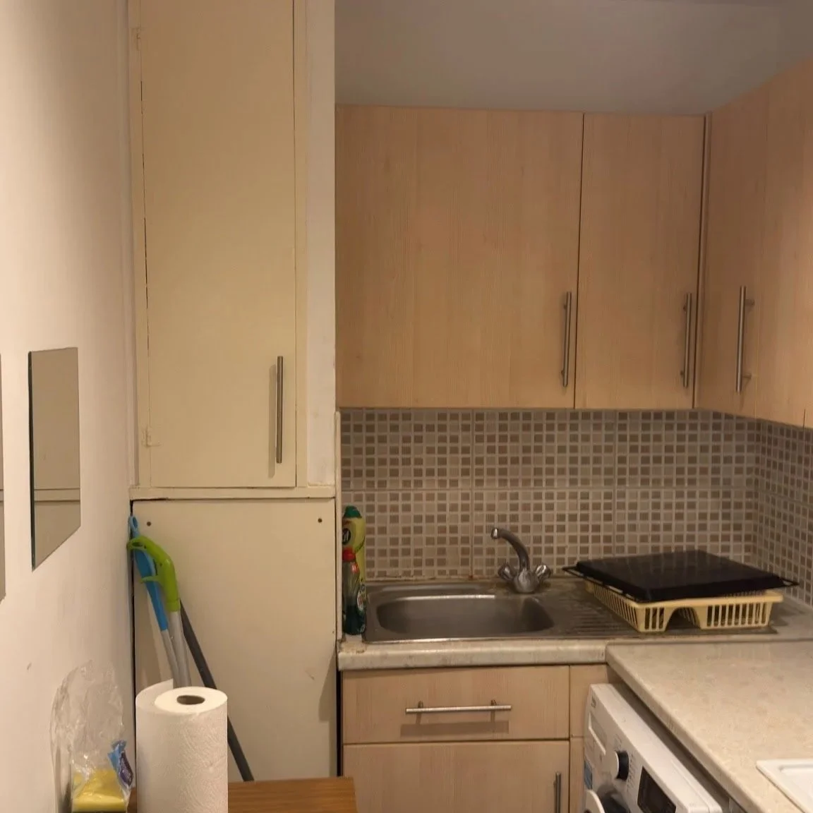 A small kitchen with beige cabinets, a tiled backsplash, a stainless steel sink, a dish drying rack, cleaning supplies near the sink, a washing machine, paper towels, a cutting board, and an electric cooktop.