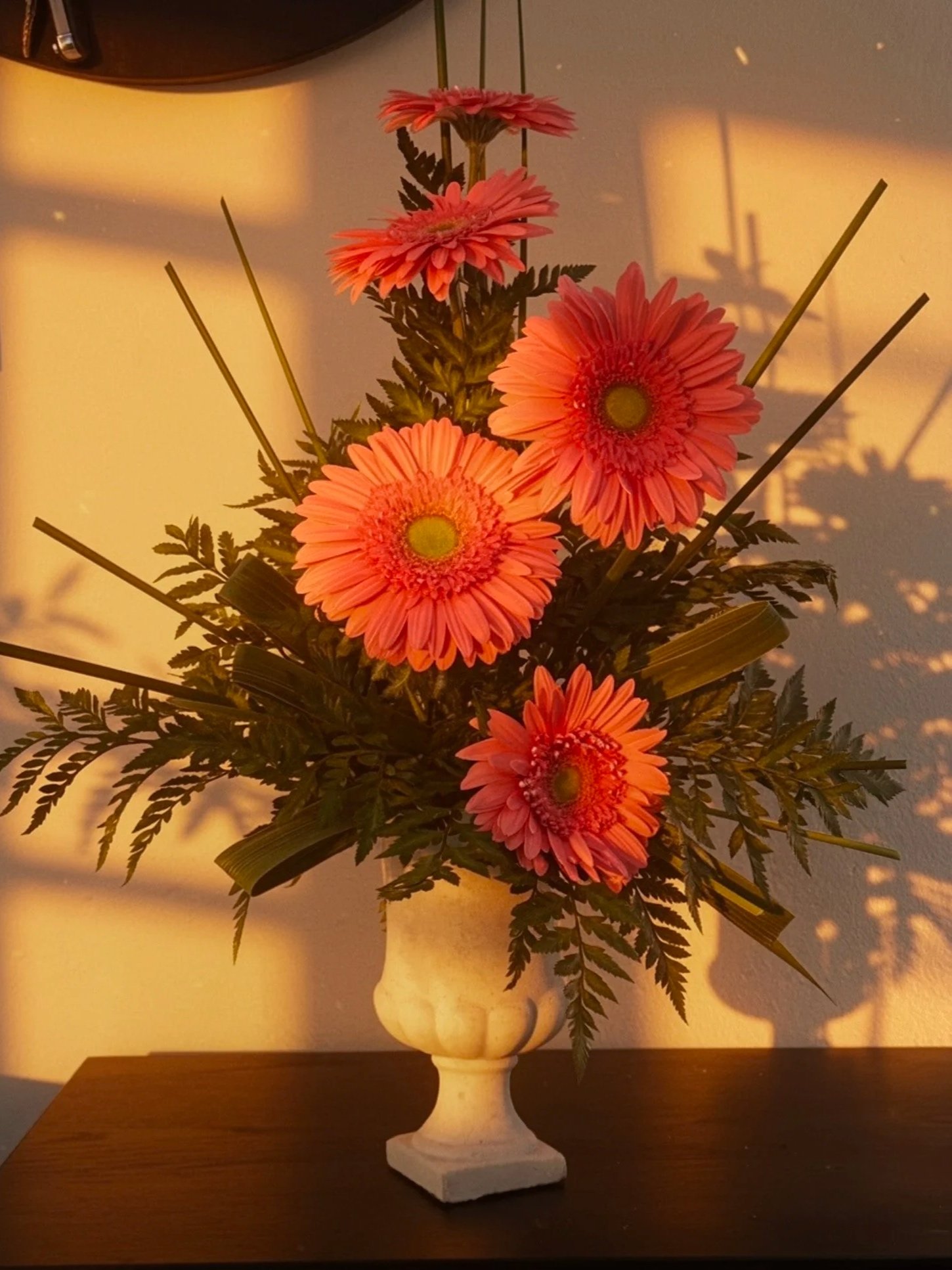 A white ceramic vase with pink gerbera daisies and greenery, placed on a wooden surface with warm lighting casting shadows.