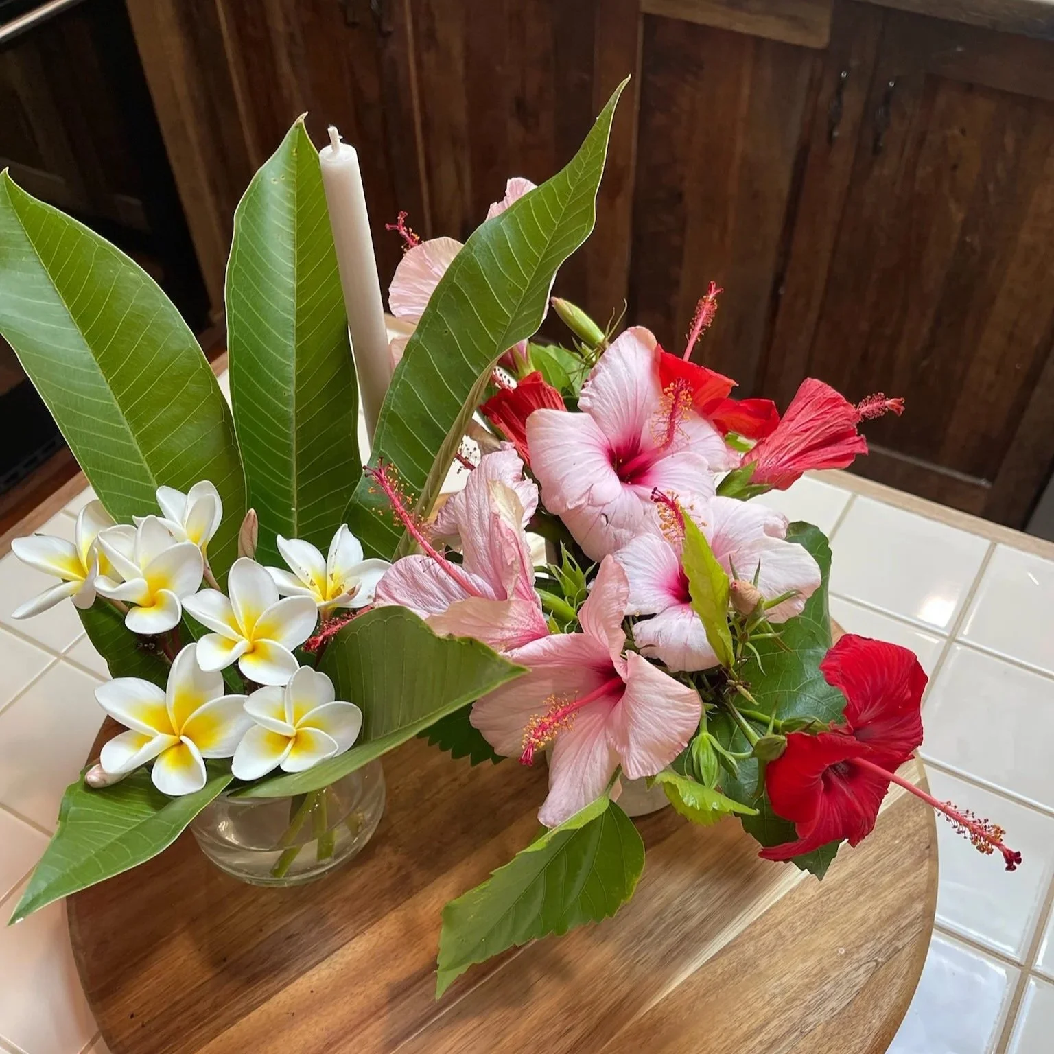 A floral arrangement in a glass vase with pink, red, and white flowers, green leaves, and a white candle on a kitchen island.
