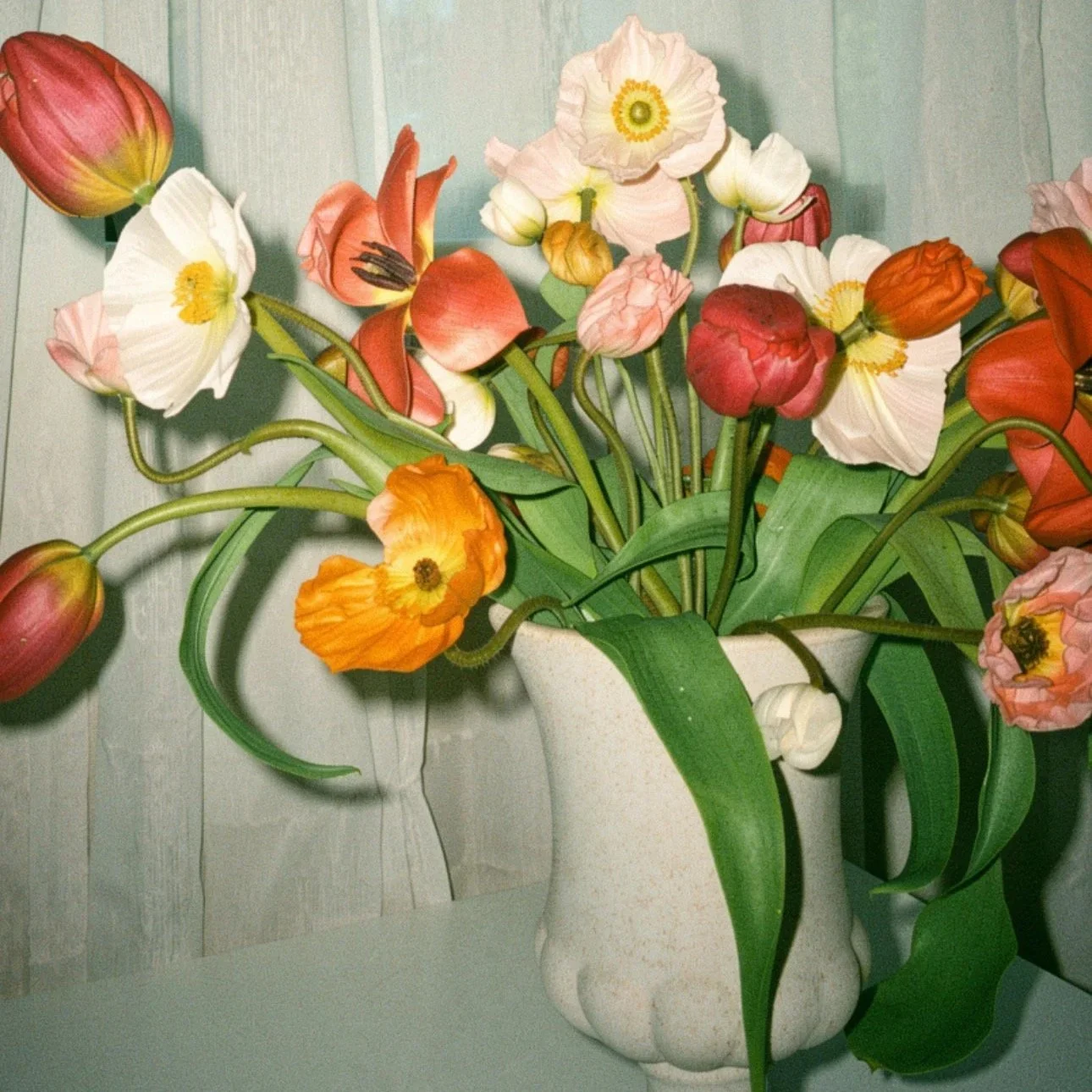 A white ceramic vase filled with pink, orange, red, and white tulips with green leaves, set against a linen background.
