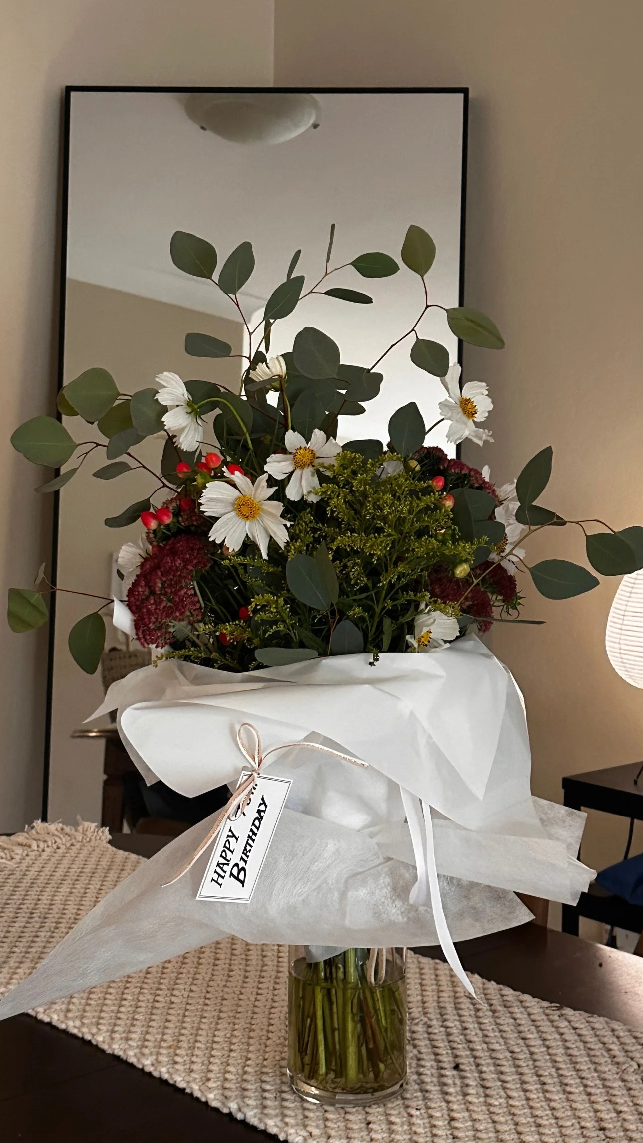 A bouquet of flowers in a clear glass vase with green stems, wrapped in white paper with a "Happy Birthday" tag. 