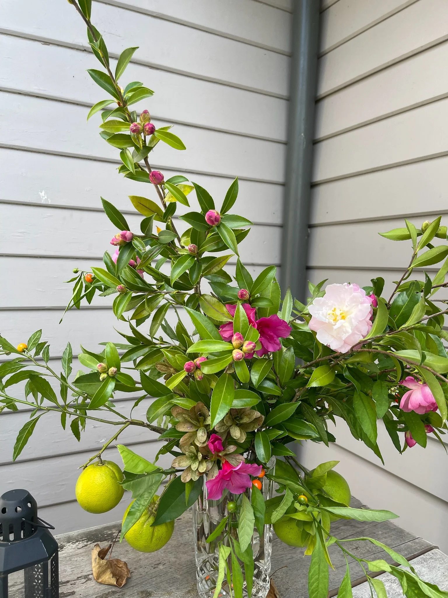 A bouquet of flowers and greenery with pink and white blossoms and two green lemons in a glass vase. 