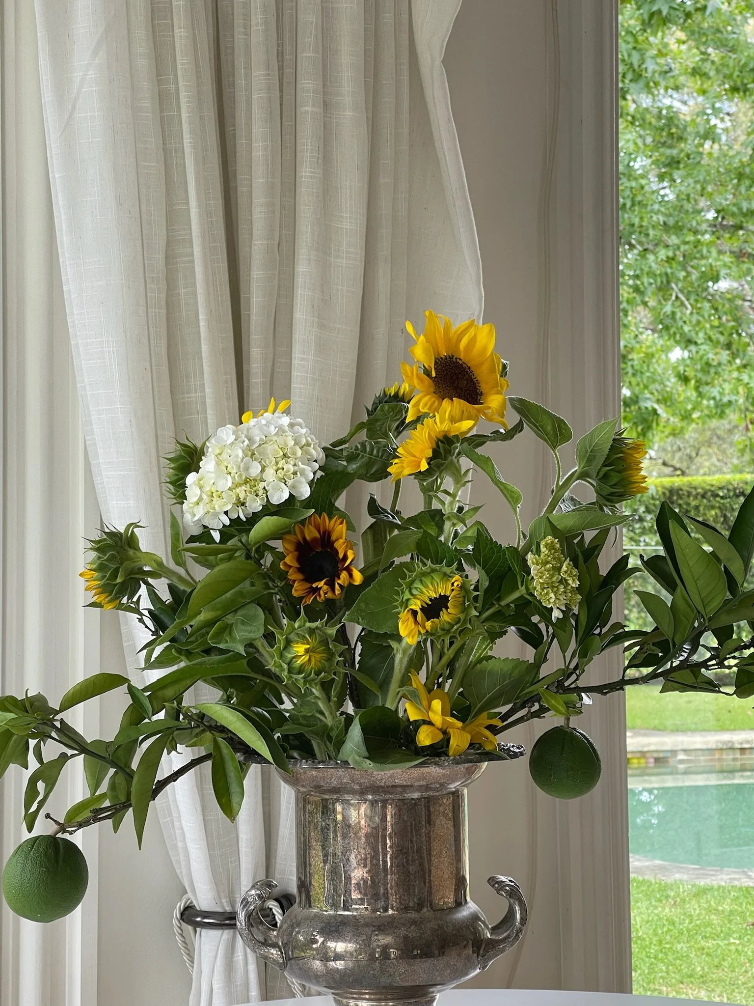 A floral arrangement with sunflowers, white hydrangeas, and green foliage in a silver urn.