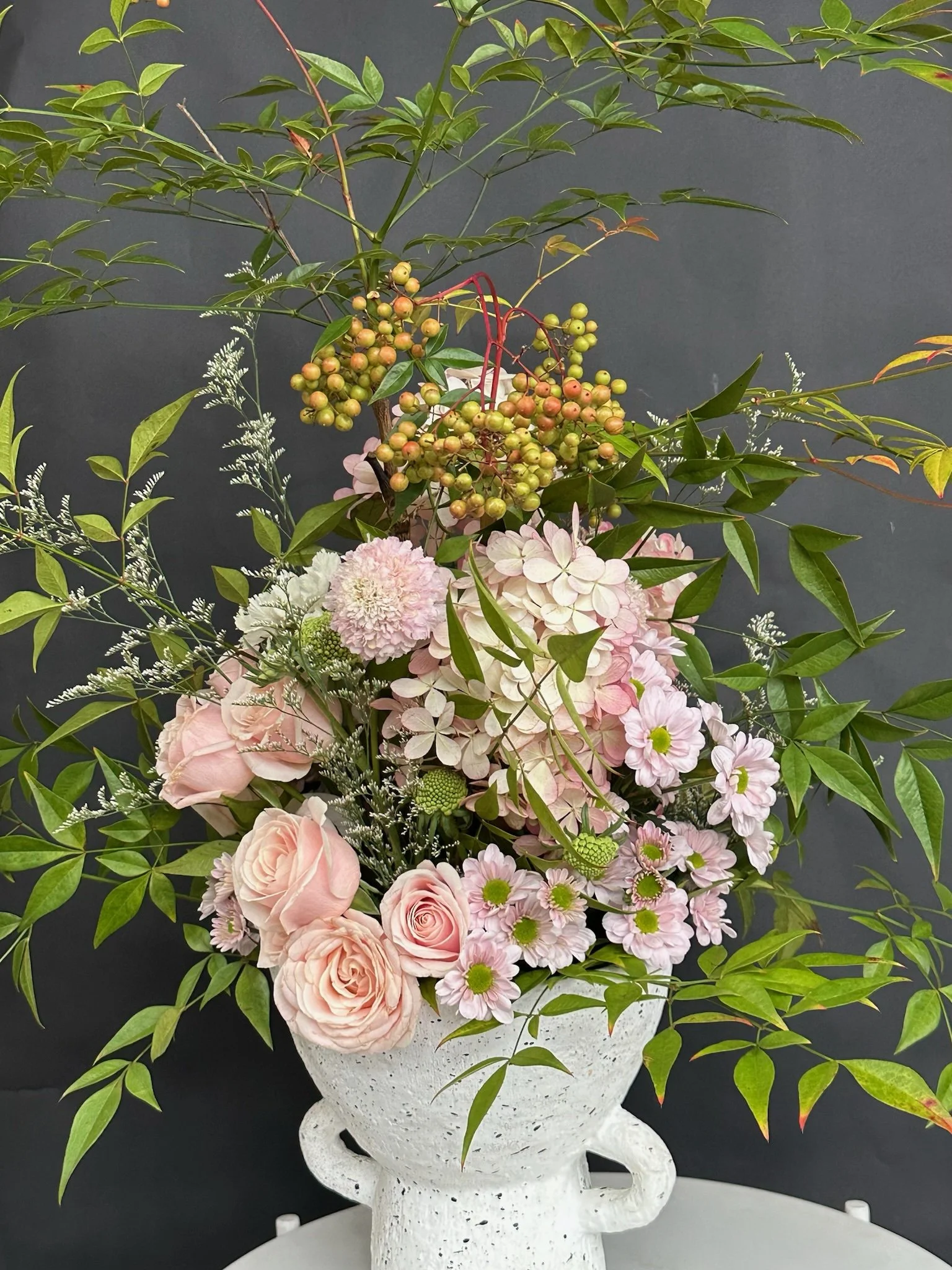A floral arrangement in a white decorative vase with swan-shaped handles, containing pink roses, white and pink hydrangeas, green foliage, and berries.