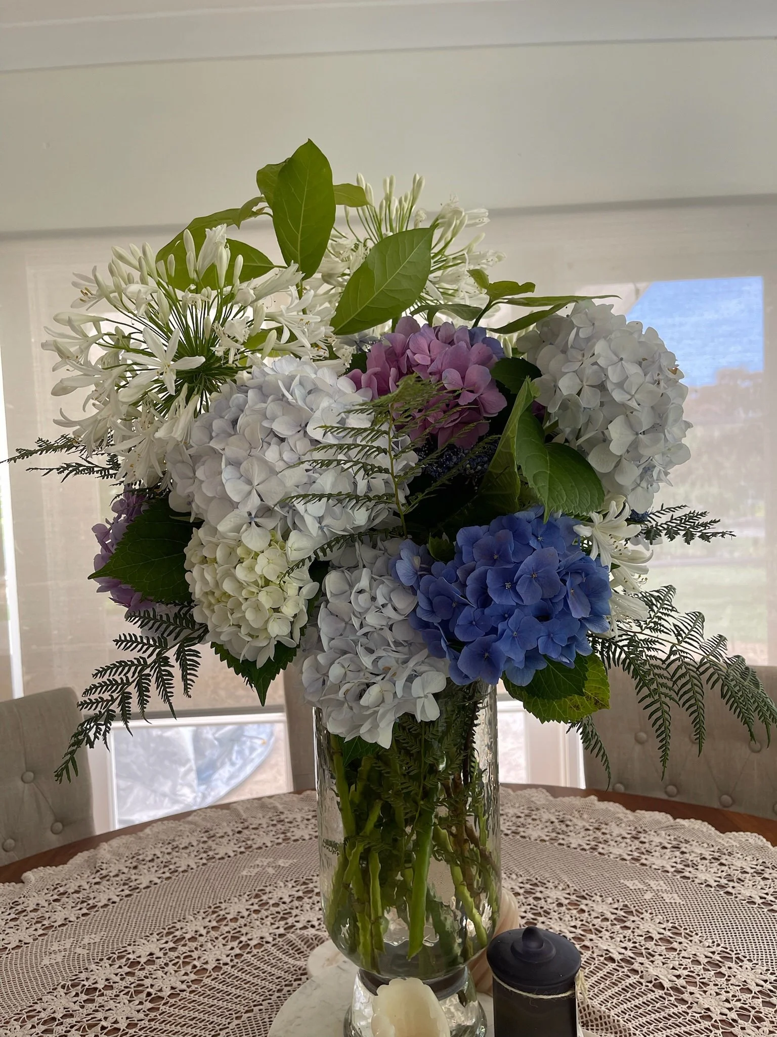 A tall glass vase filled with a large arrangement of white, purple, and blue hydrangeas, white lilies, green leaves, and ferns, placed on a table with a lace tablecloth in a room with large windows.