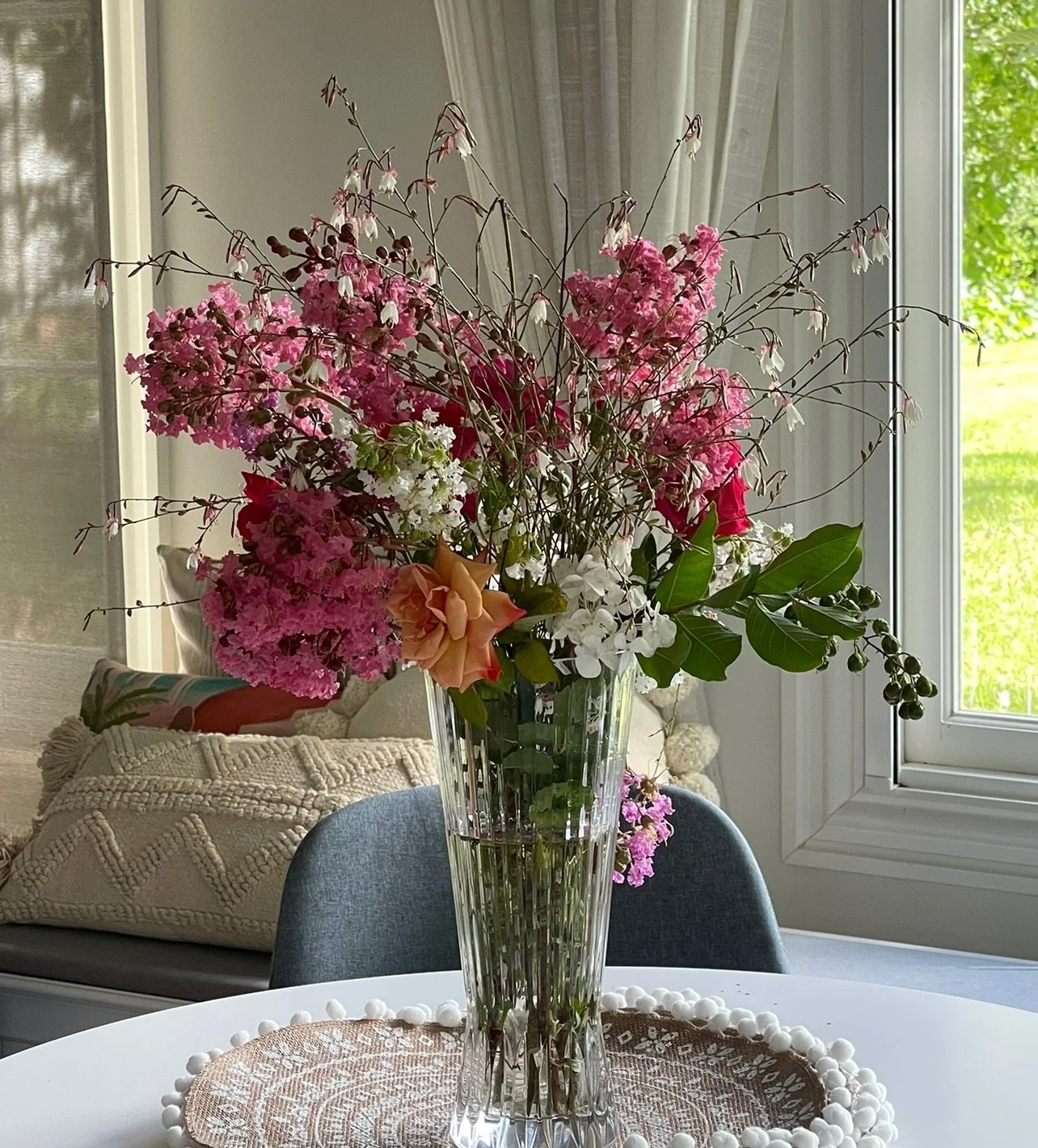 A tall glass vase filled with a colorful arrangement of pink, white, and peach flowers on a white table, with a lace doily underneath.