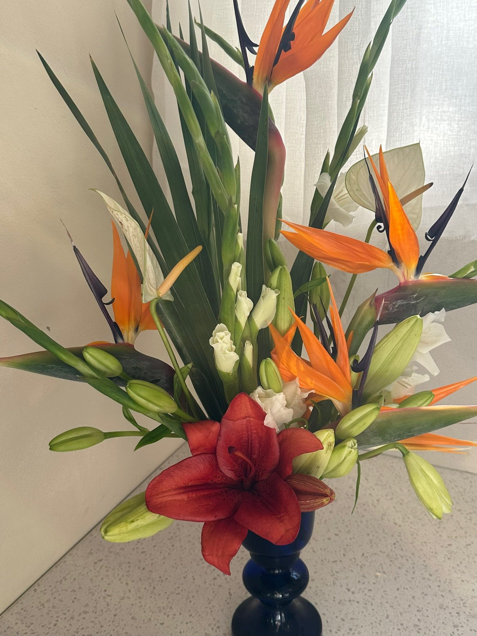 A colorful floral arrangement in a dark blue vase, featuring orange lilies, white calla lilies, white roses, red and orange anthuriums, white ginger, and green foliage.