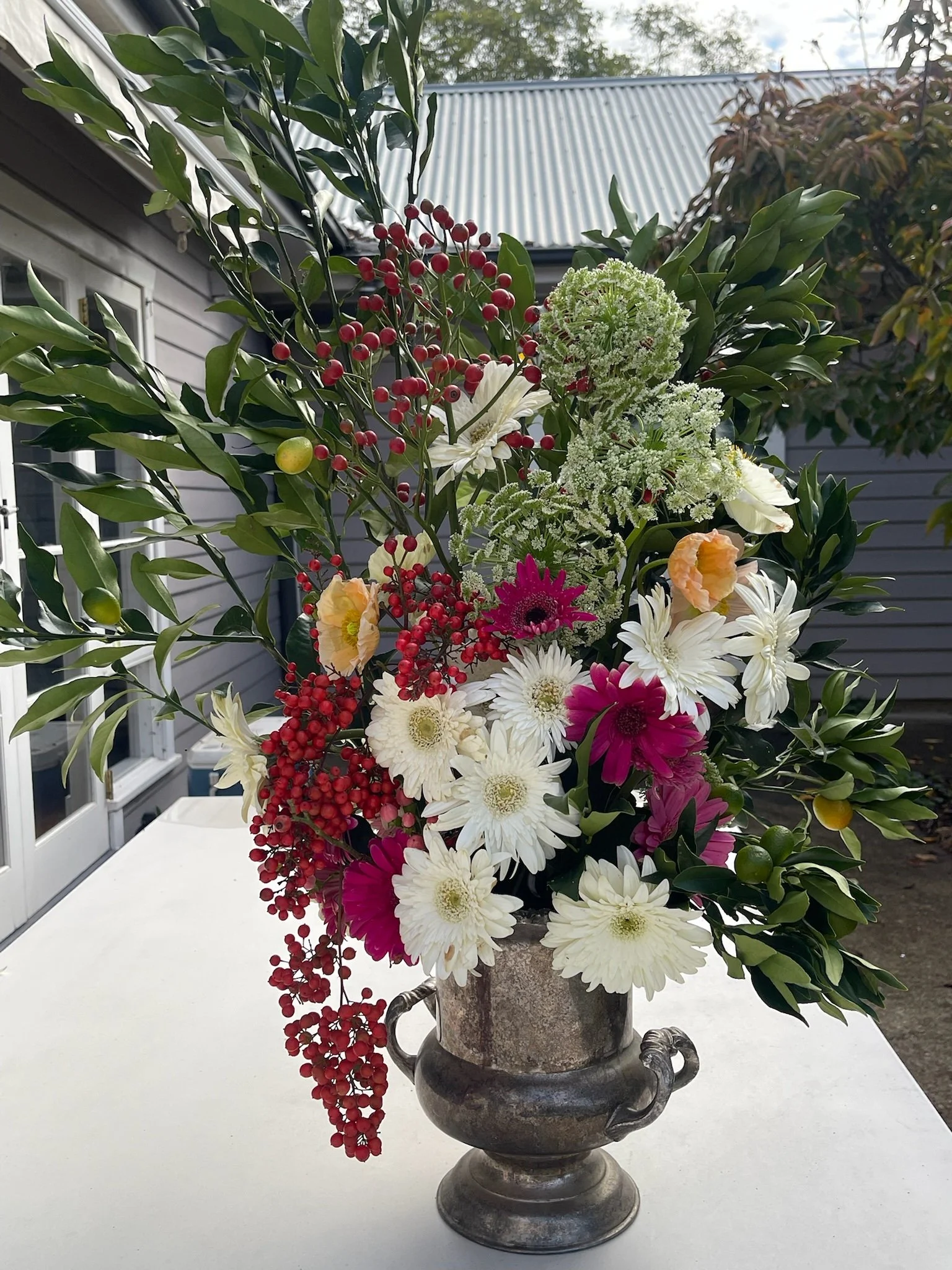 A floral arrangement in a decorative metal urn with red berries, white, pink, and yellow flowers, and green foliage, placed on a white table outdoors with a house and trees in the background.