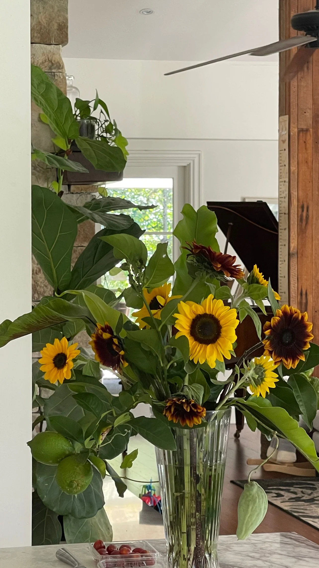 A glass vase with a sunflower bouquet, with a piano and window in the background.