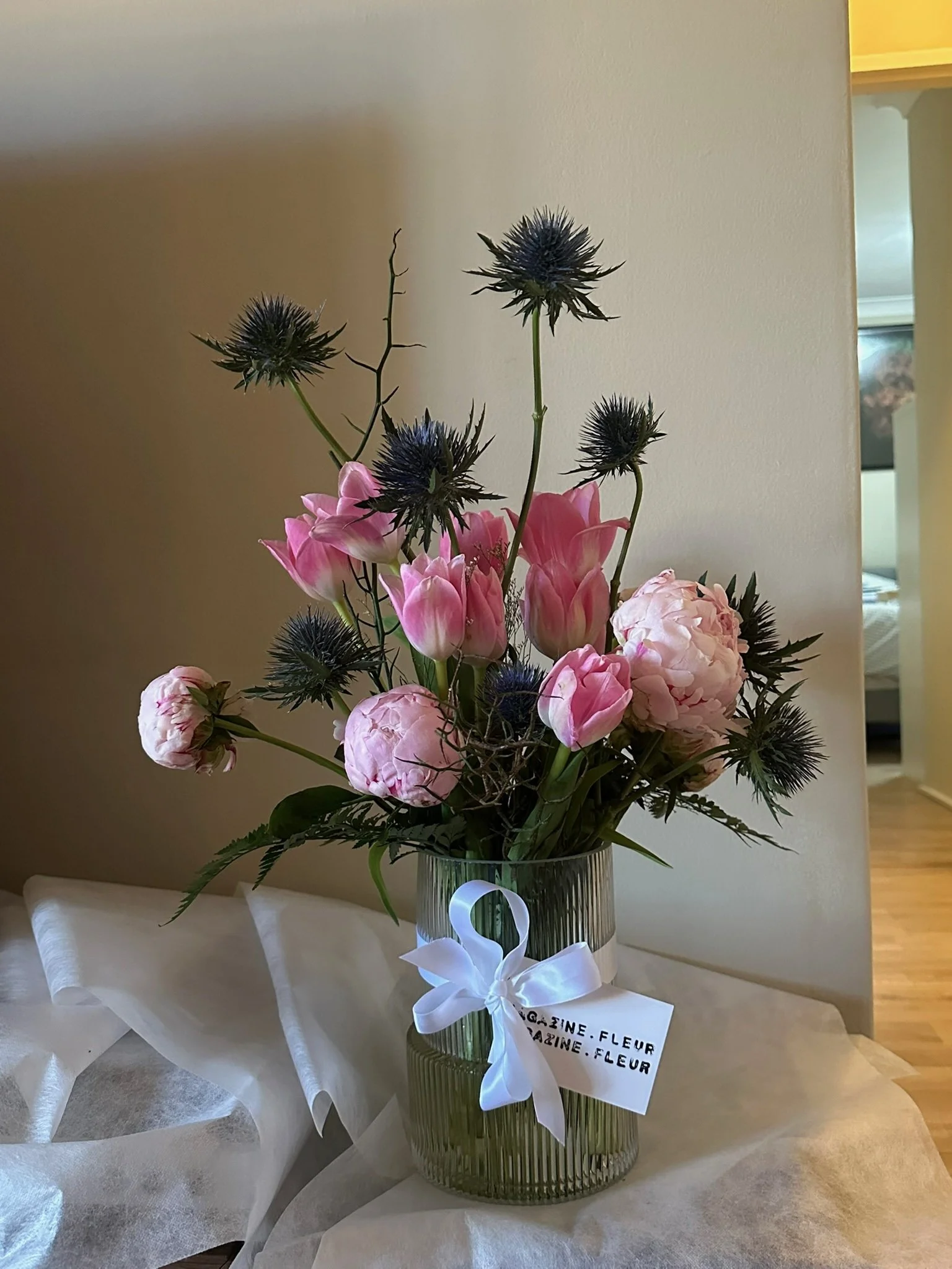 A floral arrangement in a dark green glass vase on a white table with a ribbon and tag. The flowers include pink tulips, pink peonies, and dark purple thistle flowers and ferns.