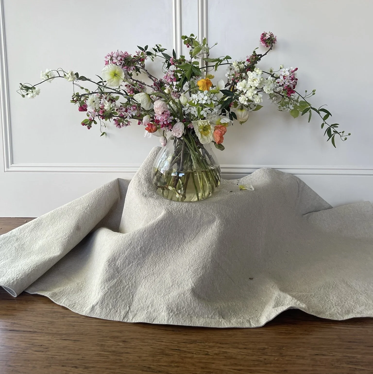 A glass vase filled with a large bouquet of mixed flowers, with a beige cloth underneath.